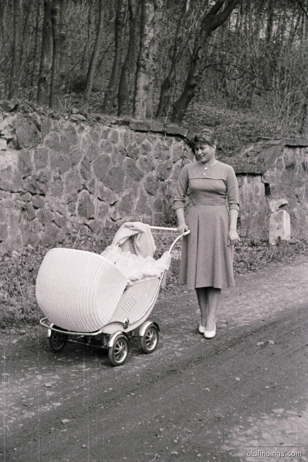 A woman walks along a paved path, pushing an elaborate wicker baby carriage. She wears a modest, knee-length dress and appears well-groomed. The background features a stone wall and trees, suggesting a rural or estate setting. Likely 1950s or 1960s, mid-century domestic scene.