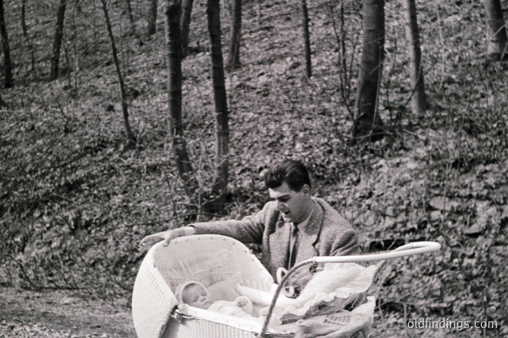 A man in a suit pushes a wicker baby carriage through a wooded area with fallen leaves. The infant within is sleeping. Likely a mid-century portrait, showcasing domestic life and early parenthood. Simple elegance & natural setting create a tranquil moment.