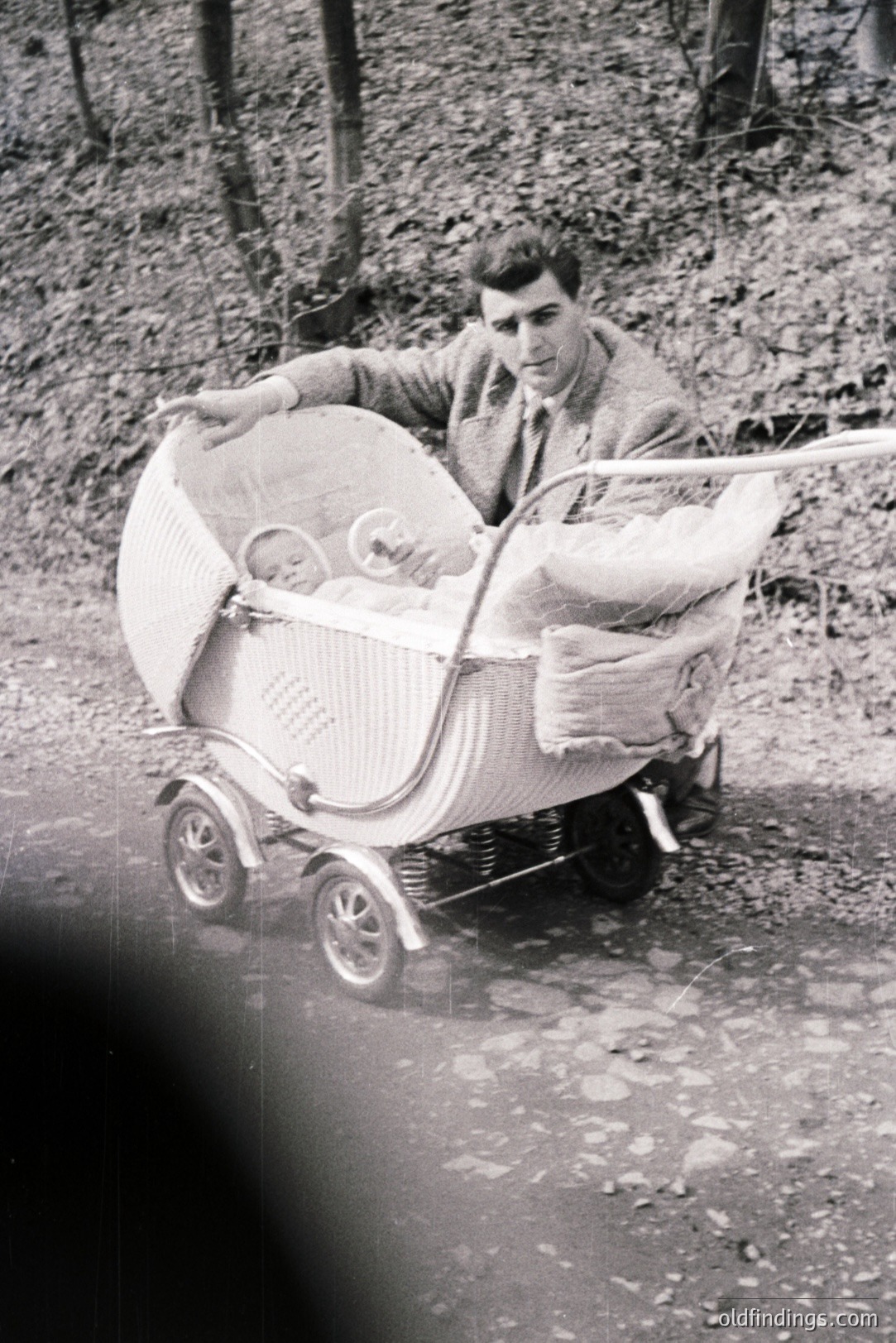 A man in a suit pushes a vintage wicker baby carriage along a wooded path. The carriage features chrome detailing and appears well-maintained. A baby, bundled in blankets, rests inside. Likely a family portrait from the 1960s or 70s, offering nostalgic appeal.