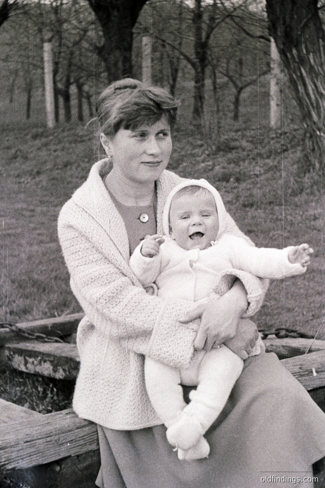 A woman in a crocheted cardigan and dress holds a baby wearing a matching outfit and bonnet. They sit on a stone bench in an outdoor setting with trees in the background. Likely a family portrait, indicative of mid-century style and domestic life. c. 1960s.