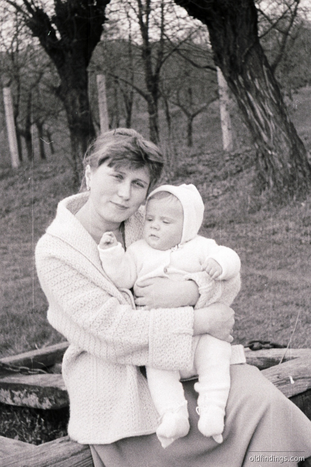 A woman holds an infant, both wearing matching knitted suits with hoods. Seated on a wooden structure near a gently sloping, treed landscape. The scene suggests a casual family portrait, likely from the 1960s or 1970s. Image shows signs of age and wear.
