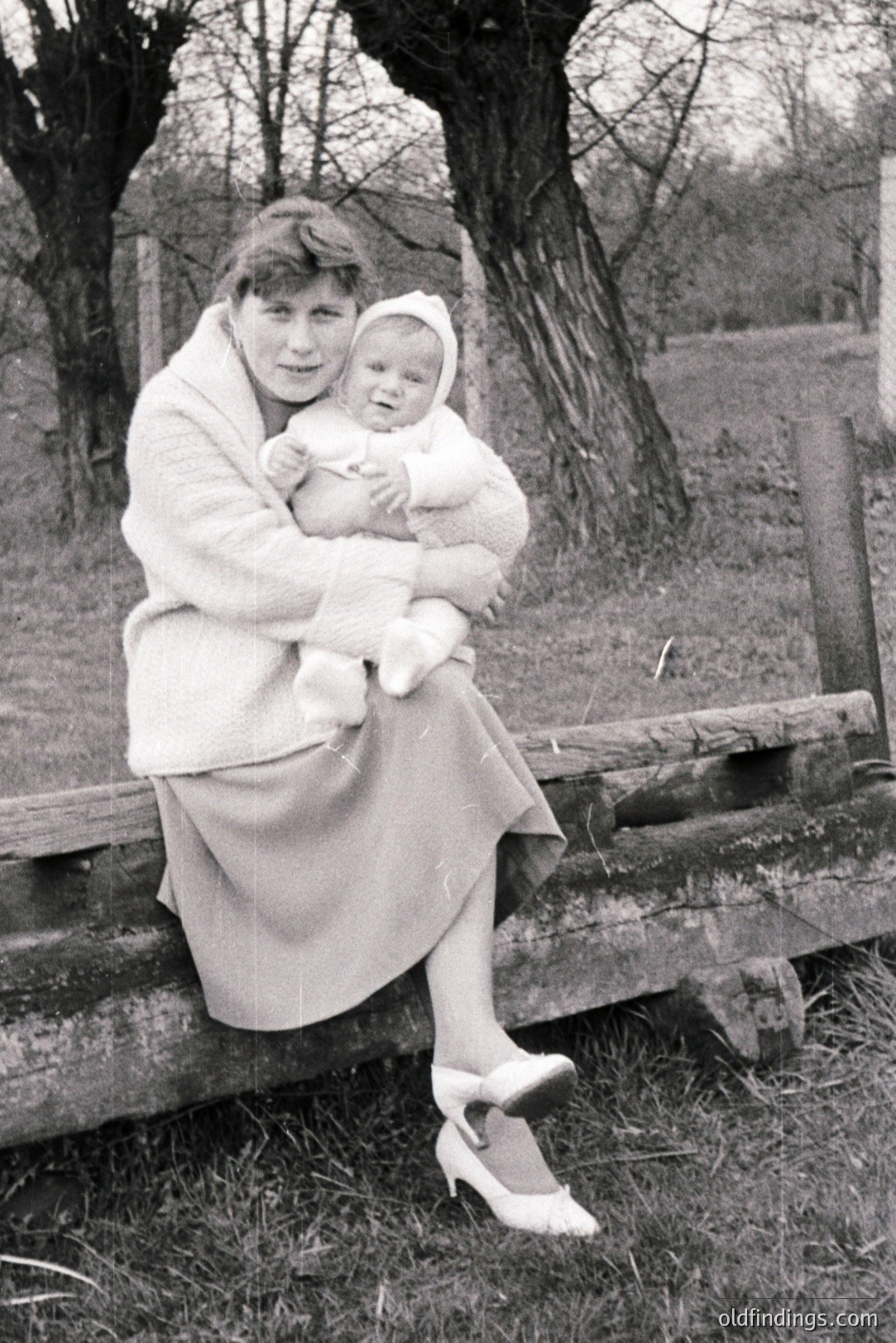 A woman in a knee-length skirt and heels cradles a baby bundled in a knit outfit. Seated on a rustic wooden barrier, they are posed against a backdrop of leafless trees and a grassy field. The image exhibits characteristics of candid family photography, likely from the 1960s or 1970s. Represents a period of modest style & familial documentation.