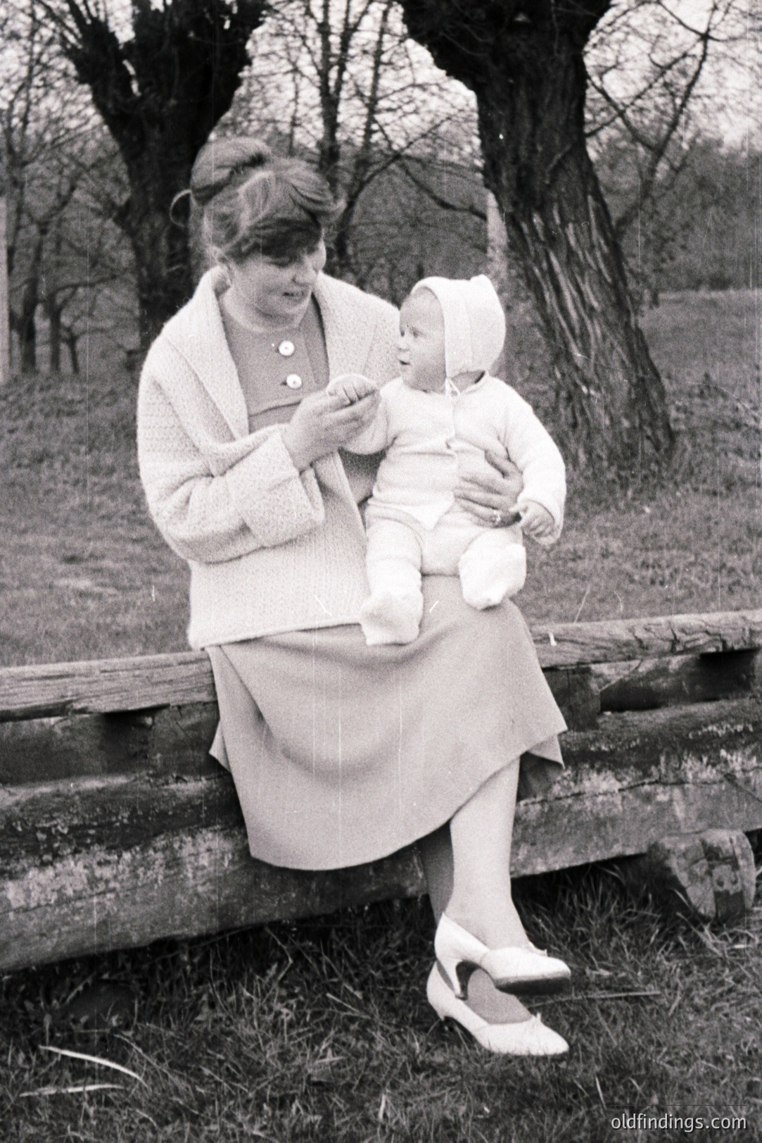 A woman sits on a wooden fence, holding a baby bundled in white. The woman wears a fitted dress suit and pointed heels, indicative of the 1960s style. Likely a family snapshot capturing a moment of maternal care outdoors. The rustic fence and simple background suggest a rural setting.