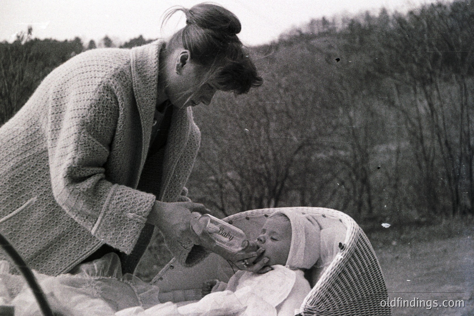A woman in a cardigan gently feeds a baby in a wicker basket outdoors. The setting appears to be a garden or park with a blurred natural backdrop. Likely a family portrait from the 1960s or 1970s. Nostalgic scene of early childcare. Potentially useful for vintage design or historical research.