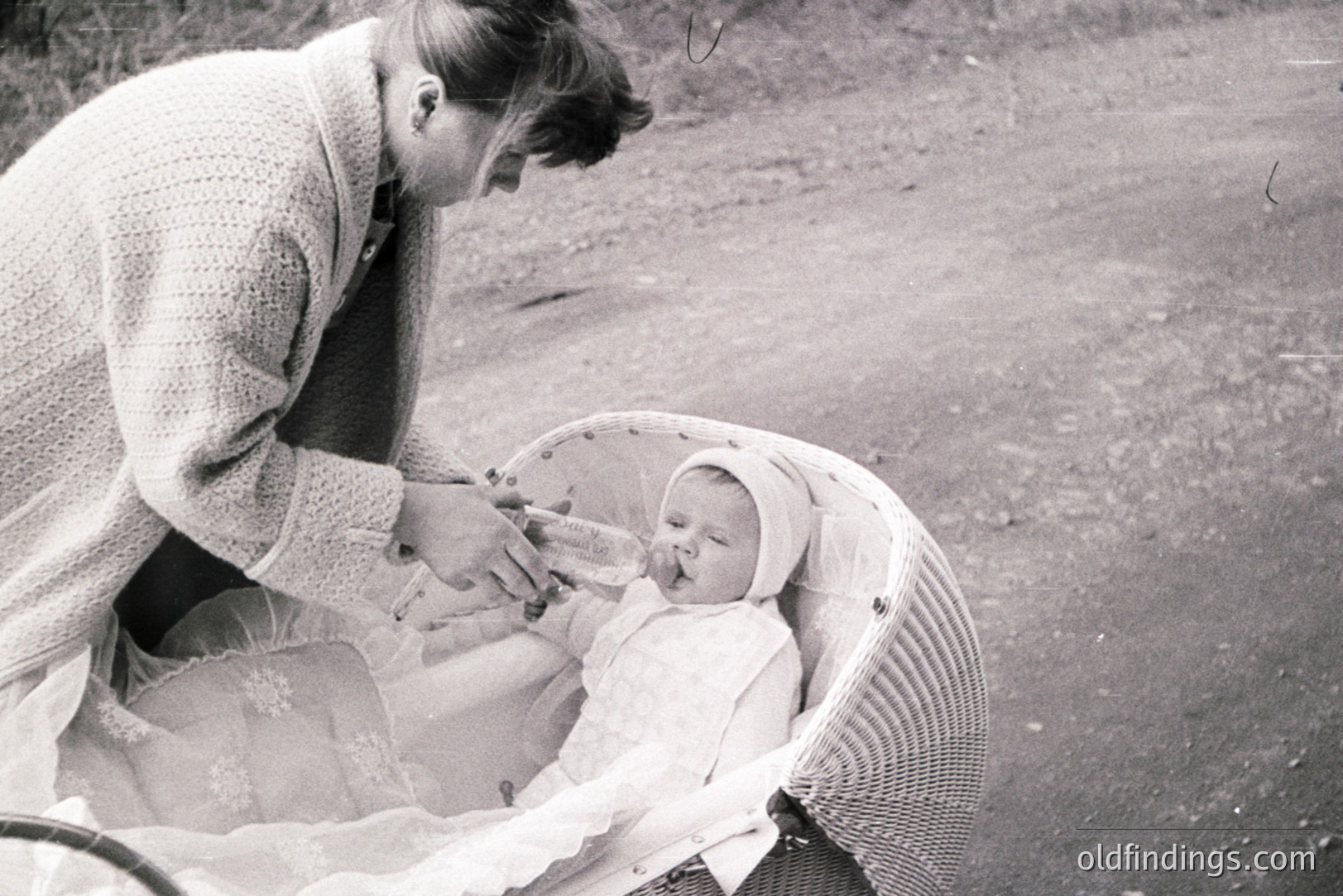 A woman in a knitted sweater tends to an infant nestled in a wicker carriage, likely outdoors. The baby wears a bonnet and lace-trimmed clothing. The image evokes a domestic, intimate moment, possibly from the 1960s or 70s. Style suggests family documentation. Potential for sentimental stock use.
