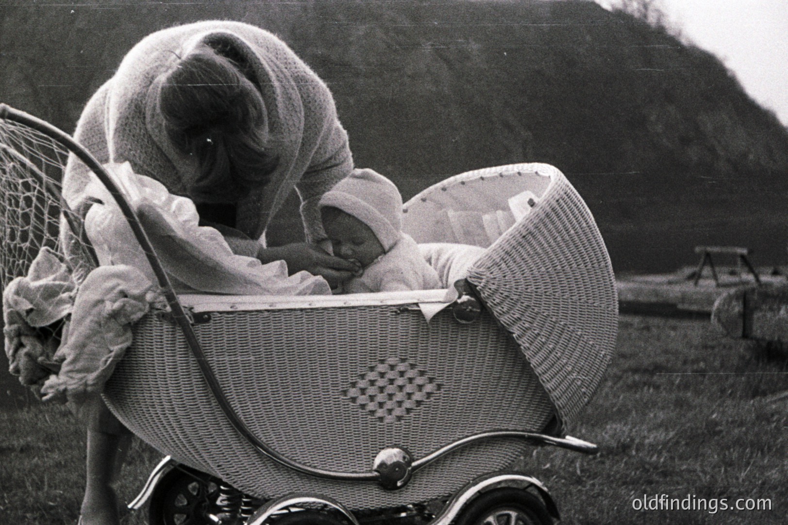 A young baby rests in an elaborate wicker pram, likely mid-century. The mother, bundled in a large sweater, leans over the infant. Grassy field backdrop with a picnic table visible. A glimpse of a nostalgic, domestic moment.