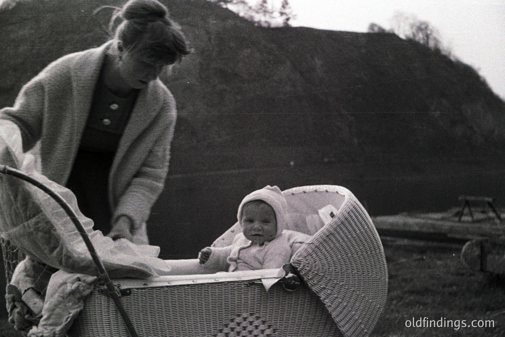 A young mother stands near a wicker baby carriage containing an infant in a bonnet, posed outdoors on a slight incline. The style of dress and carriage suggests a mid-20th century domestic scene. The backdrop features a grassy hillside. A potential reference for vintage advertising.