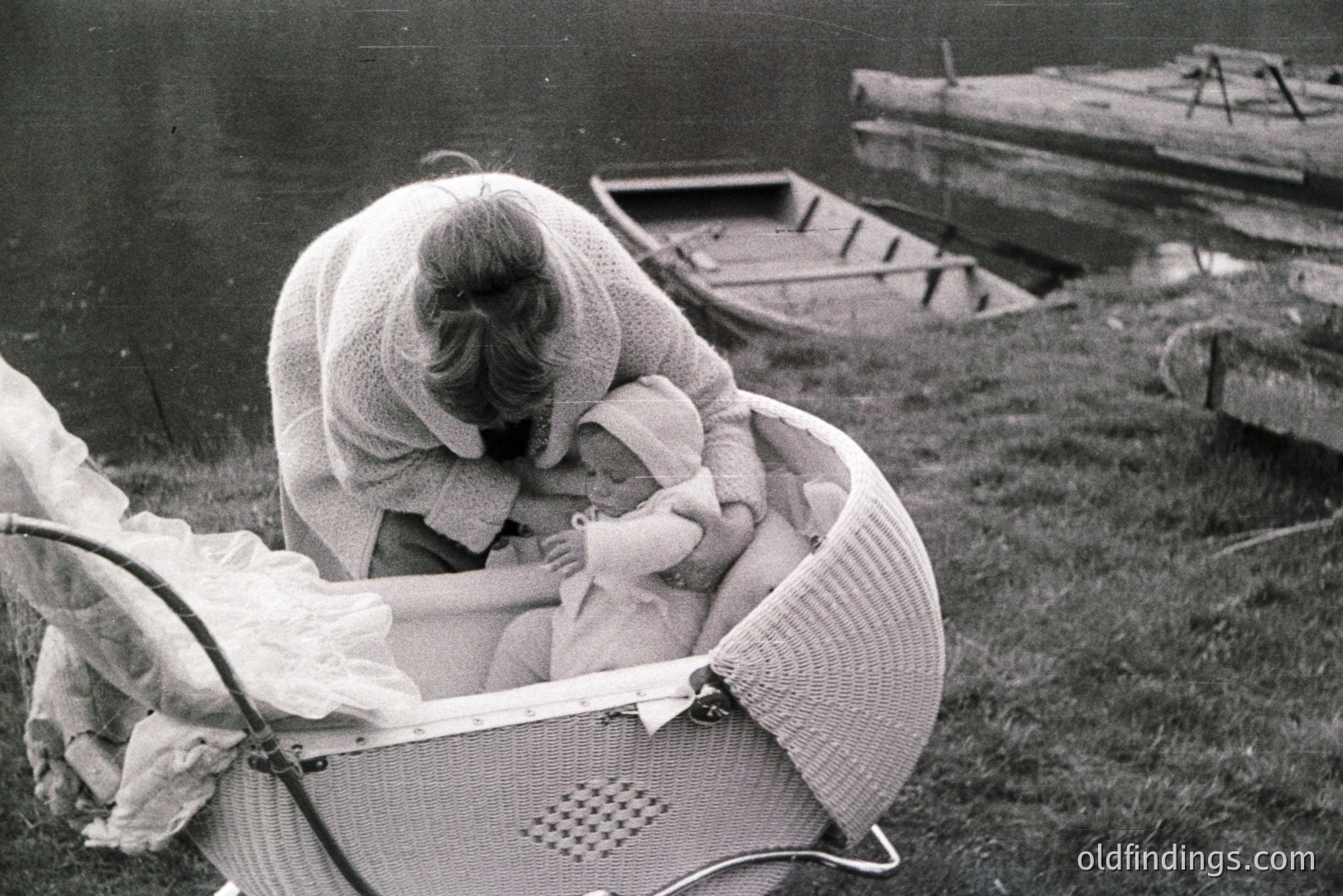 A woman in a crocheted shawl tends to an infant nestled in an ornate, woven wicker crib beside a body of water. The scene evokes a sense of domestic tranquility. Likely 1960s, the photograph’s composition suggests a lifestyle study or family portrait.