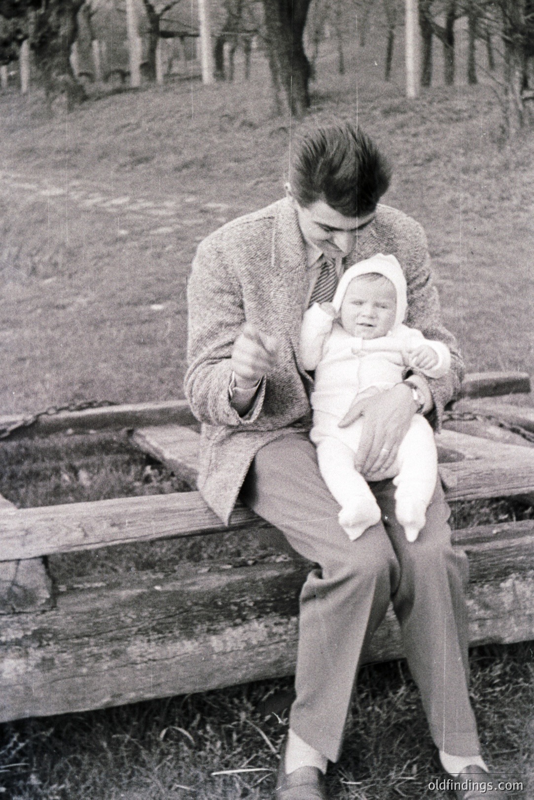 A man in a tweed jacket and tie cradles an infant dressed in white. Seated on a weathered wooden fence, they appear outdoors with a grassy hillside visible in the background. Likely a family snapshot from the mid-20th century. Potential use for vintage family or baby-related stock.
