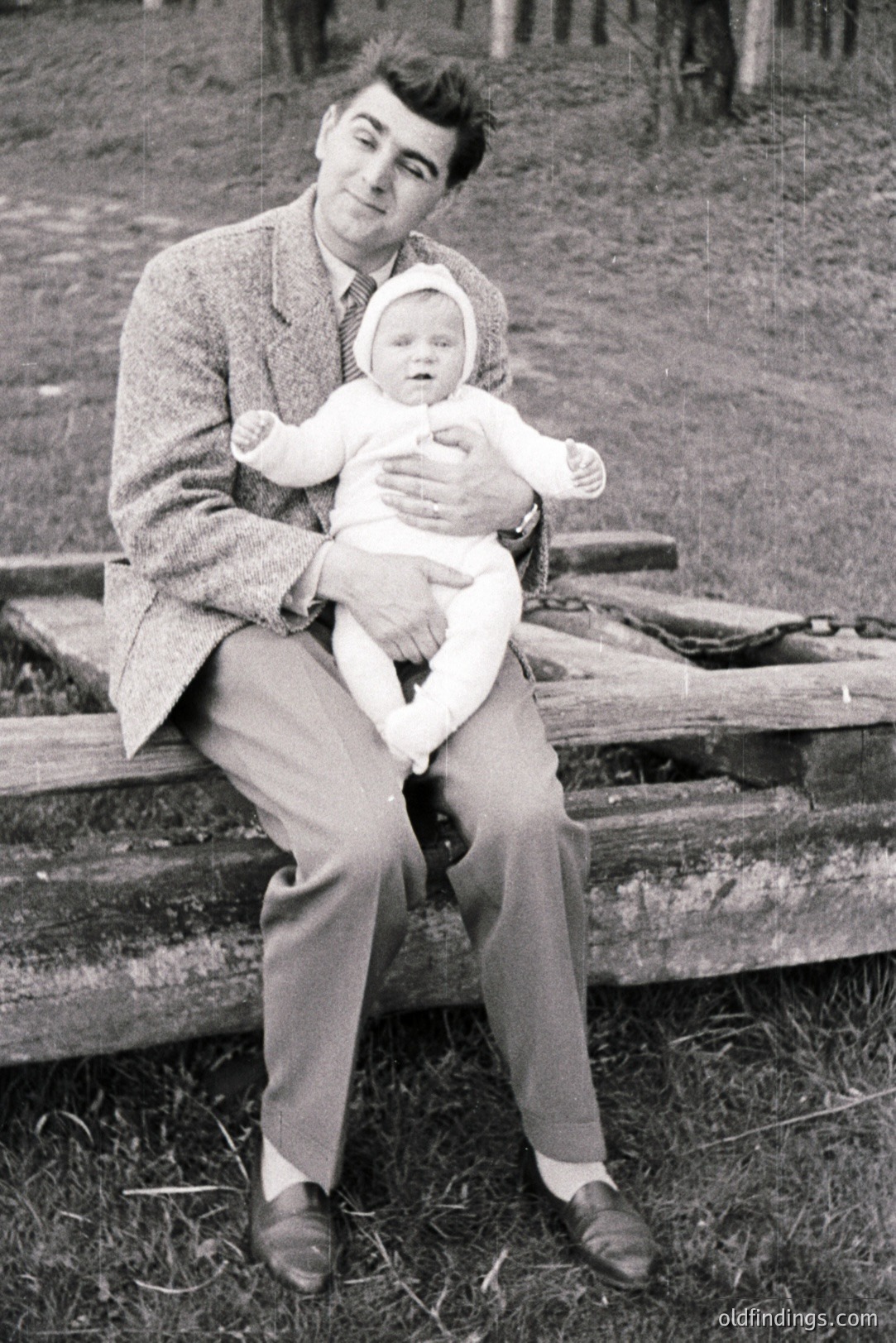 Man in a tweed jacket and trousers cradles a baby wrapped in white, seated on a wooden structure, possibly a bench. Appears to be an outdoor portrait, likely mid-20th century due to clothing style. Simple, direct composition ideal for family portraits or archival use.