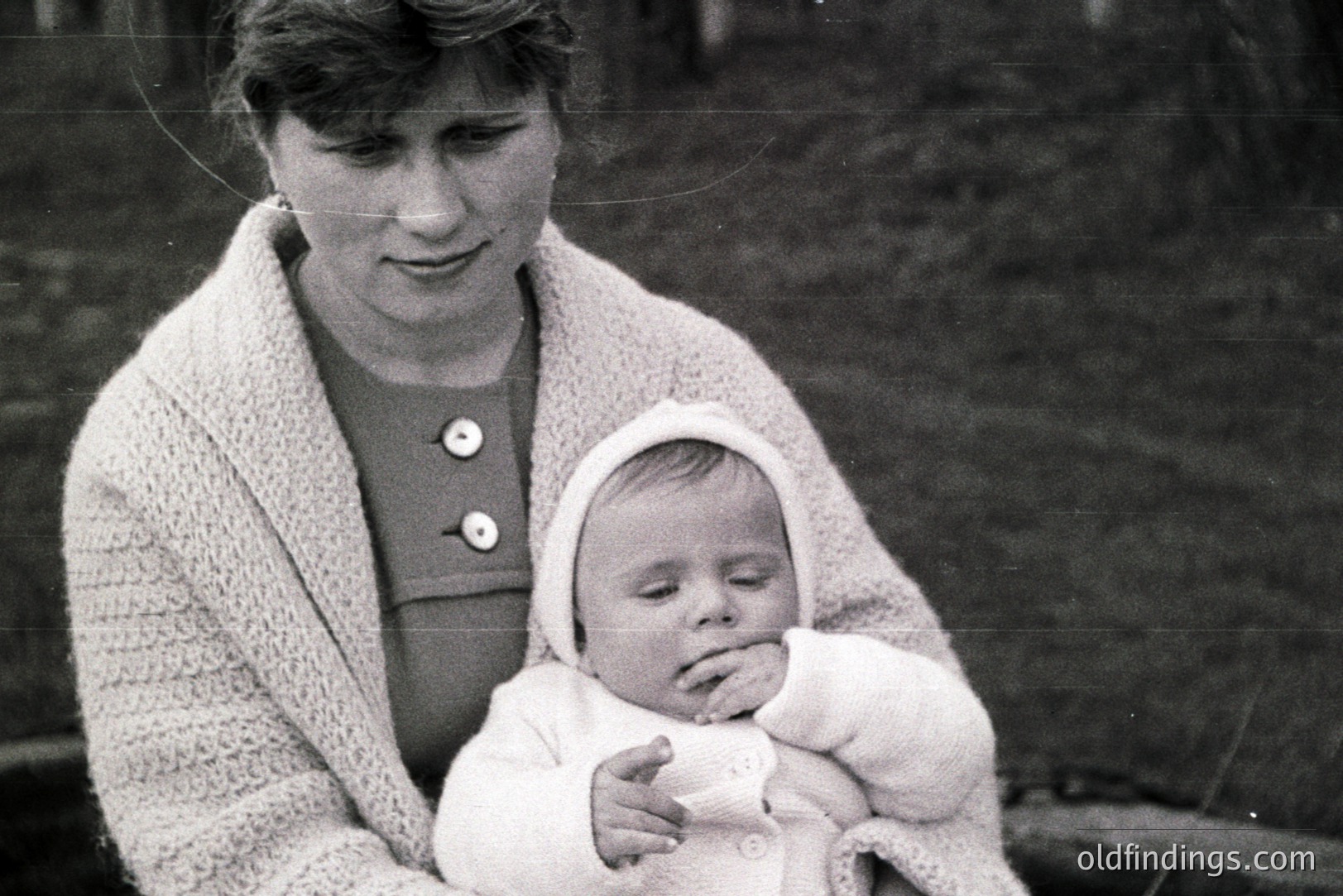 A young woman in a dark dress and knitted cardigan cradles a sleeping infant wrapped in a textured white garment. The scene appears to be outdoors, with a blurred natural background. Likely a family portrait, capturing a tender moment. Estimated 1960s-1970s. Potential design reference for vintage fashion.