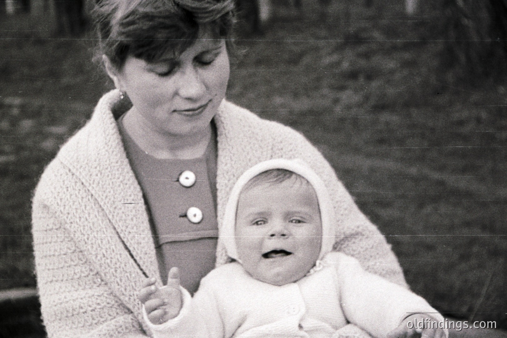 A mother and baby in a tender, intimate portrait. The mother wears a textured sweater and button-down dress, cradling an infant adorned with a matching bonnet and outfit. Appears to be a candid, informal family moment captured outdoors. Likely mid-1960s or 70s. Strong reference for vintage fashion & family imagery.