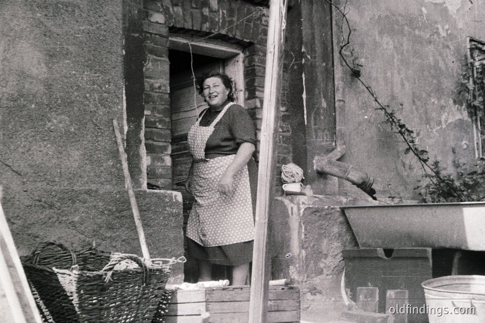 A woman stands in a weathered stone doorway, apron adorned with a patterned print. A woven basket, wooden crates, and a metal sink suggest a workspace or utility area. The rough-hewn brick and aged plaster hint at a rural or historic setting. Likely 1950s-1970s, evocative of domestic labor and traditional life.