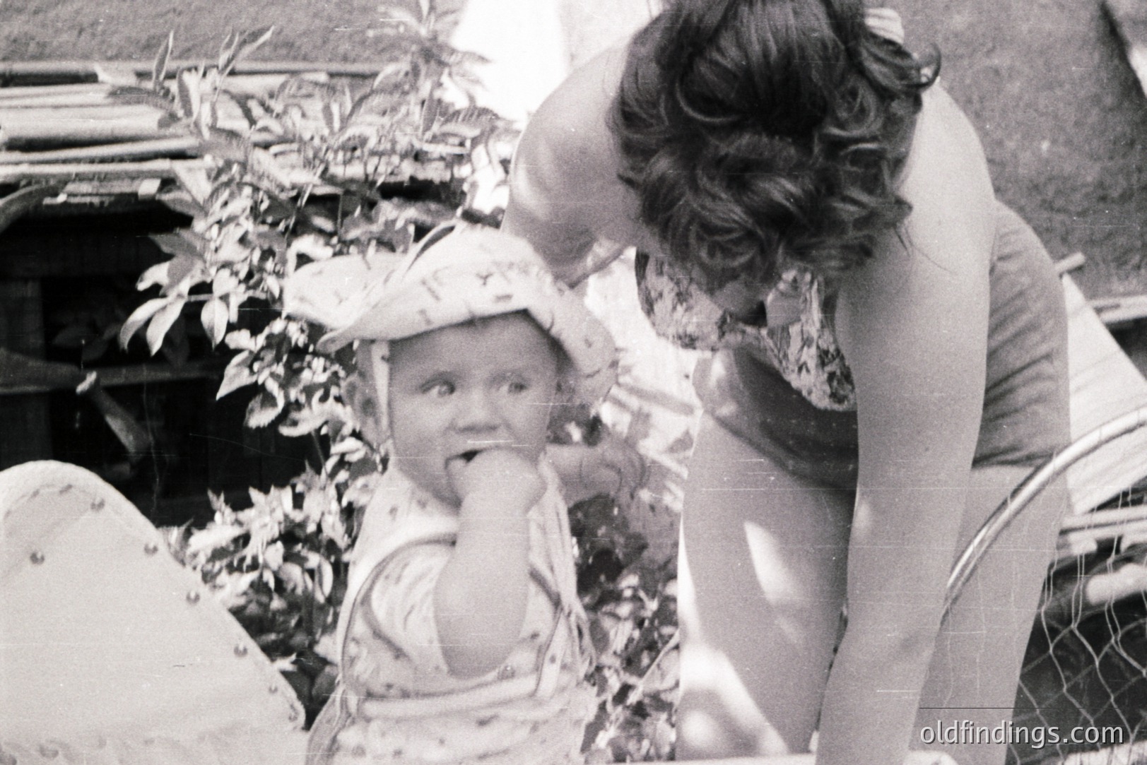 A young child with a patterned sun hat sits outdoors, appearing to eat something, while a woman leans in closely. A glimpse of garden foliage and fencing is visible. Likely a candid snapshot reflecting family life, c. 1970s. Potential stock value for nostalgic family themes.