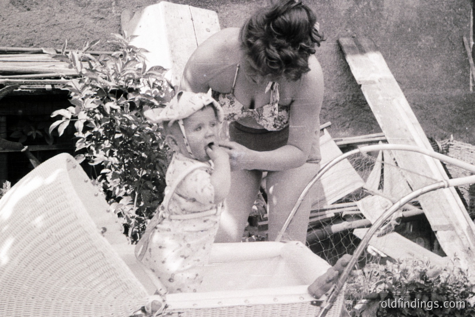 A young child sits in a wicker chair, appearing to receive a treat from a woman in a floral bathing suit. The scene is set outdoors, possibly a garden or patio, evidenced by foliage and weathered wood. Likely mid-20th century, possibly 1950s-1970s, based on the fashion and photographic style. Could be stock photography illustrating family leisure.