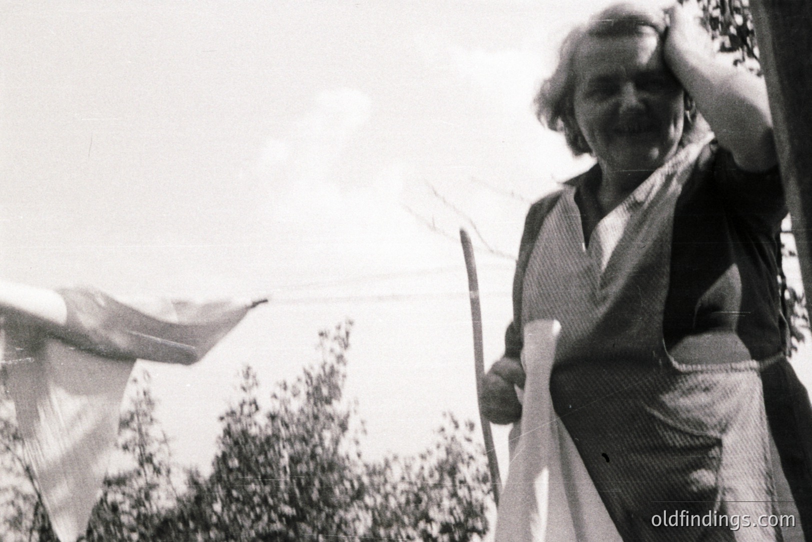 A woman in a dark dress and patterned apron hangs laundry on a line outdoors. She holds a sheet, appearing to shield her eyes from the sun. Likely a candid snapshot, possibly a domestic scene from the mid-20th century. Offers insight into everyday life.