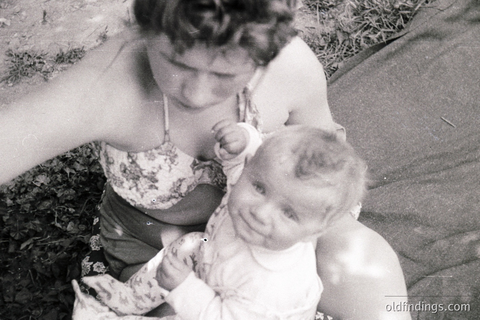 Black and white image captures a mother cradling a baby in what appears to be a garden setting. The mother wears a floral-patterned top. The baby, centrally framed, looks directly at the camera. Visible grain suggests a snapshot from the mid-20th century. Likely 1950s-1970s era.