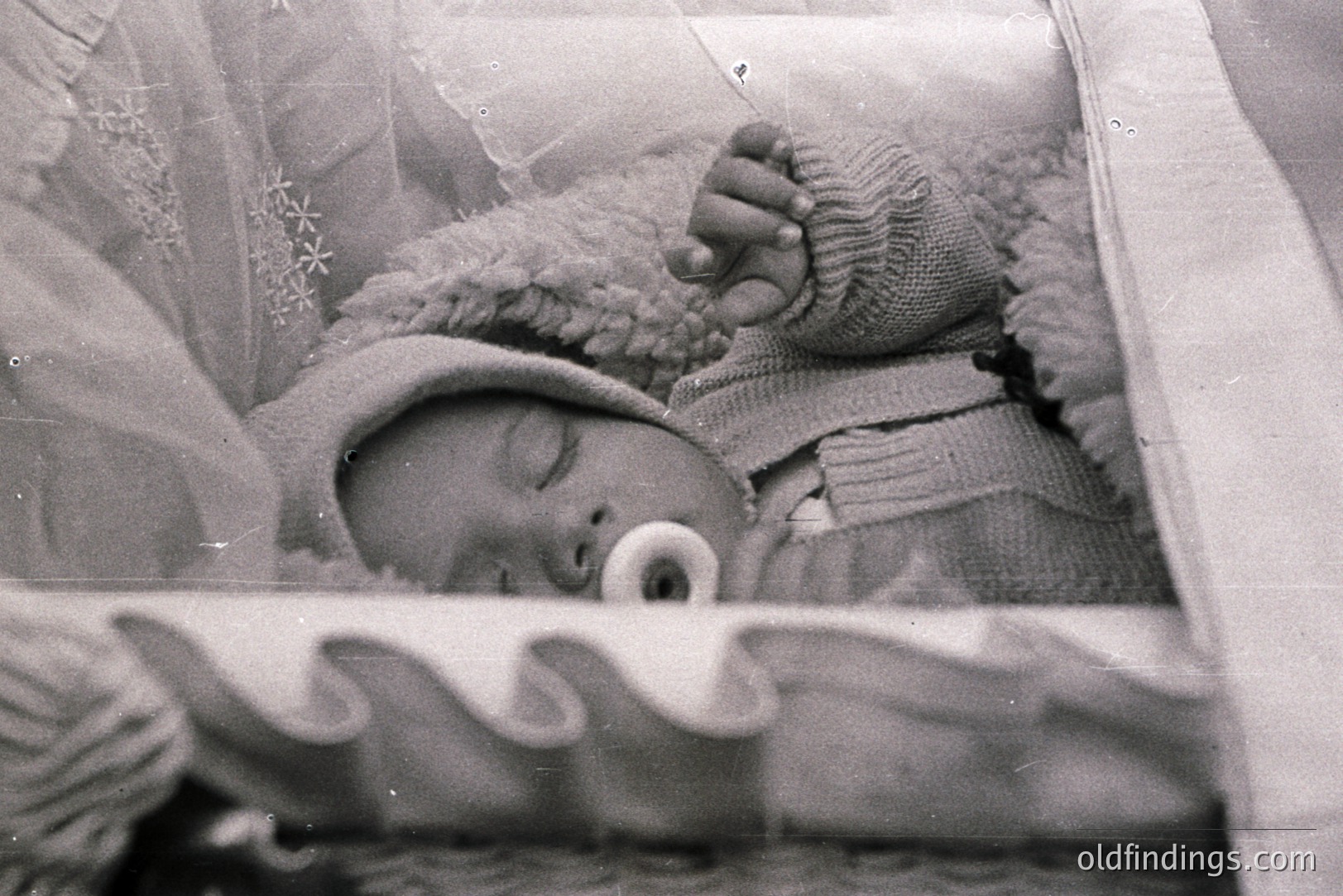 A sleeping infant, swaddled in a knitted blanket and cap, rests within a crib. The scene appears intimate, possibly a personal family photograph. Likely 1960s-70s based on photographic style and dress. A floral patterned curtain provides a soft backdrop. Gentle and poignant portrait.