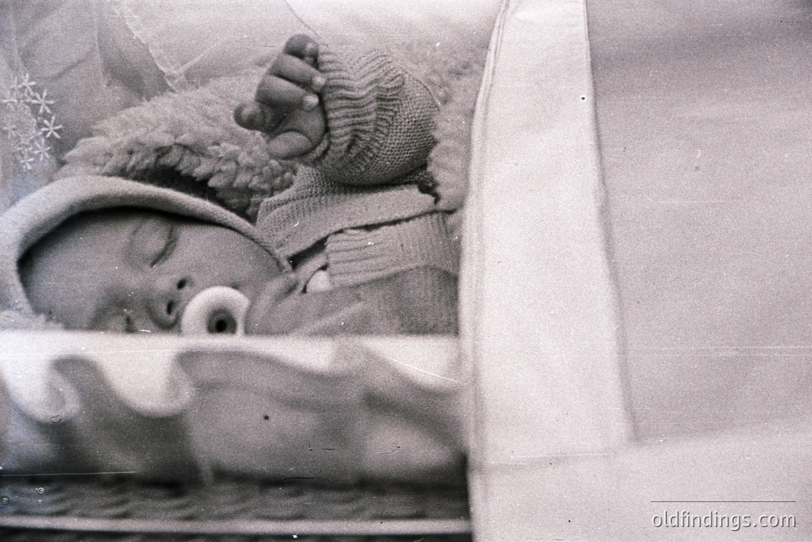Infant sleeps peacefully bundled in a knitted bonnet and textured clothing. Vintage photograph likely from the mid-20th century, exhibiting classic studio portraiture style. Crib bars are visible, suggesting a formal setting. A pacifier is visible in the child’s mouth. Likely used for family documentation or early commercial baby product advertising.
