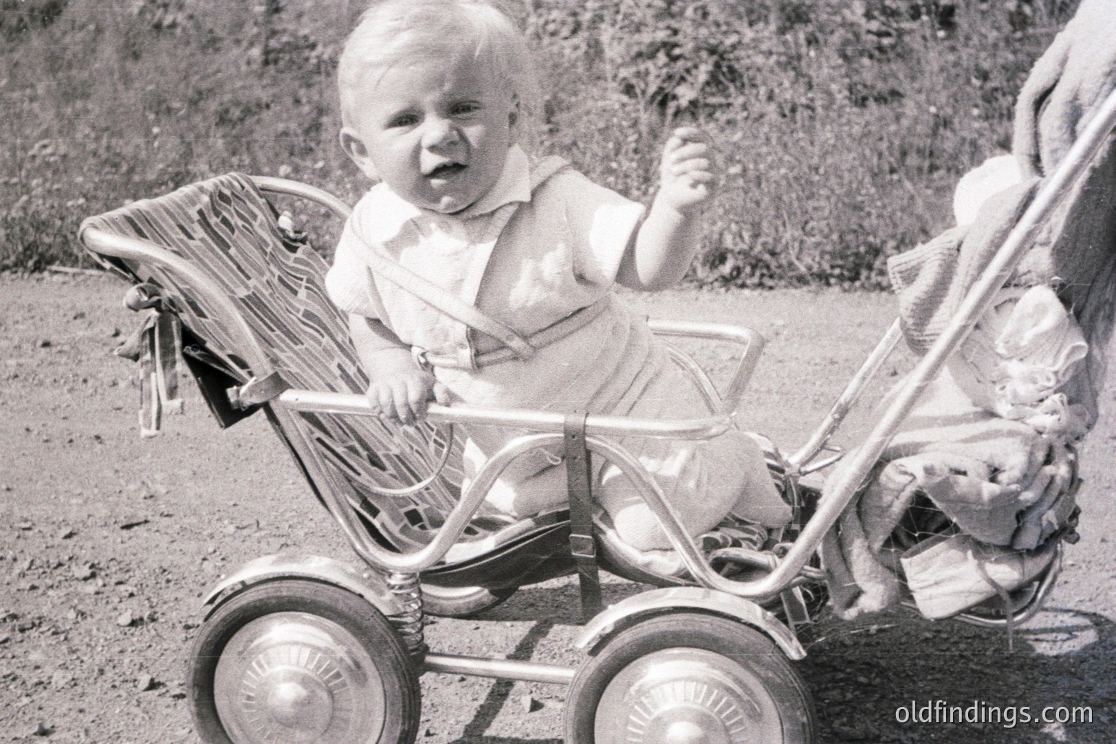 An infant sits in a vintage, wire-spoke stroller, raising one hand. The child wears a light-colored, collared outfit. Background suggests a grassy, outdoor setting. Likely a candid snapshot, indicative of domestic family life. Appears to be from the 1950s or 1960s.