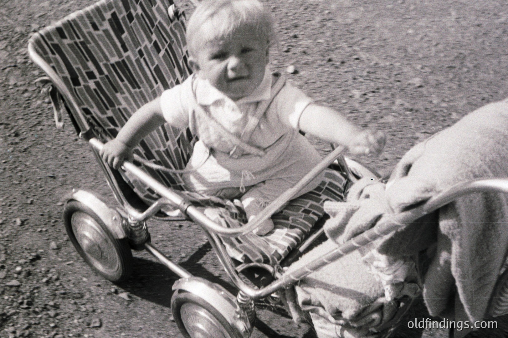 A young child sits in a vintage metal stroller, raised arm appearing animated. The stroller's design suggests a mid-20th century style. Faded blanket partially obscures the lower right corner. Likely a candid family snapshot.