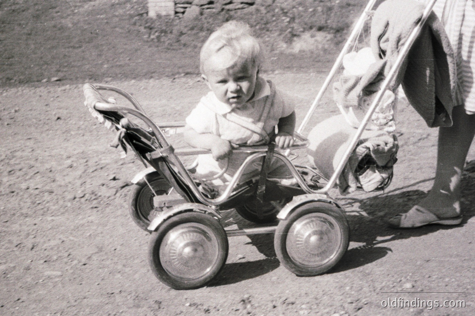 A young child sits in a vintage, chrome-framed tricycle with balloon tires. The child wears a short-sleeved shirt and suspenders. Partial view of an adult, likely a caregiver, is visible on the right. Likely 1950s-1960s, capturing a commonplace family moment. Valuable for nostalgic stock or design use.