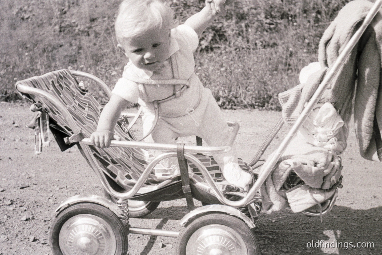 A young child, appearing to be a toddler, is playfully posed on a vintage stroller outdoors. The child is clad in a light-colored romper and has a characteristic short hairstyle of the era. The stroller exhibits a classic design with wire-spoke wheels. Likely dating to the 1950s-1960s. Excellent reference for period design or nostalgic stock imagery.