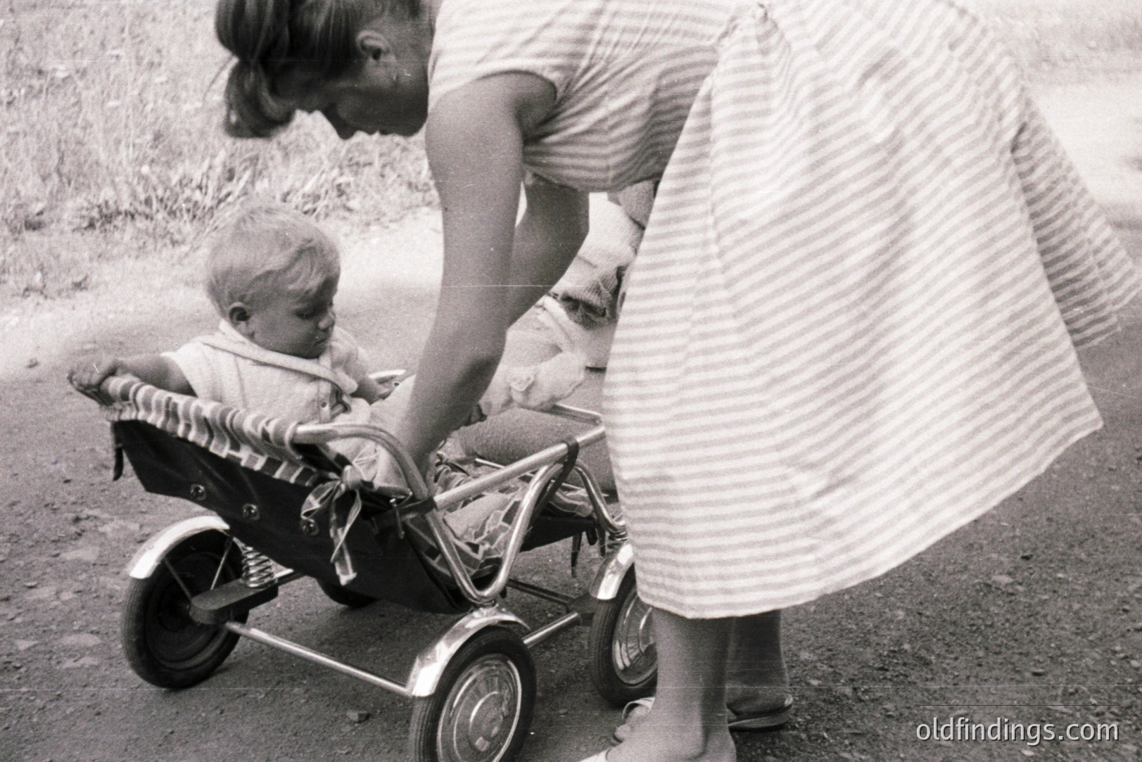 A young boy sits in a doll carriage, being gently steered by a woman in a striped dress. The scene, likely a backyard or driveway, is captured in grainy black and white. The doll carriage's design suggests a mid-20th century aesthetic. A nostalgic glimpse of childhood play.
