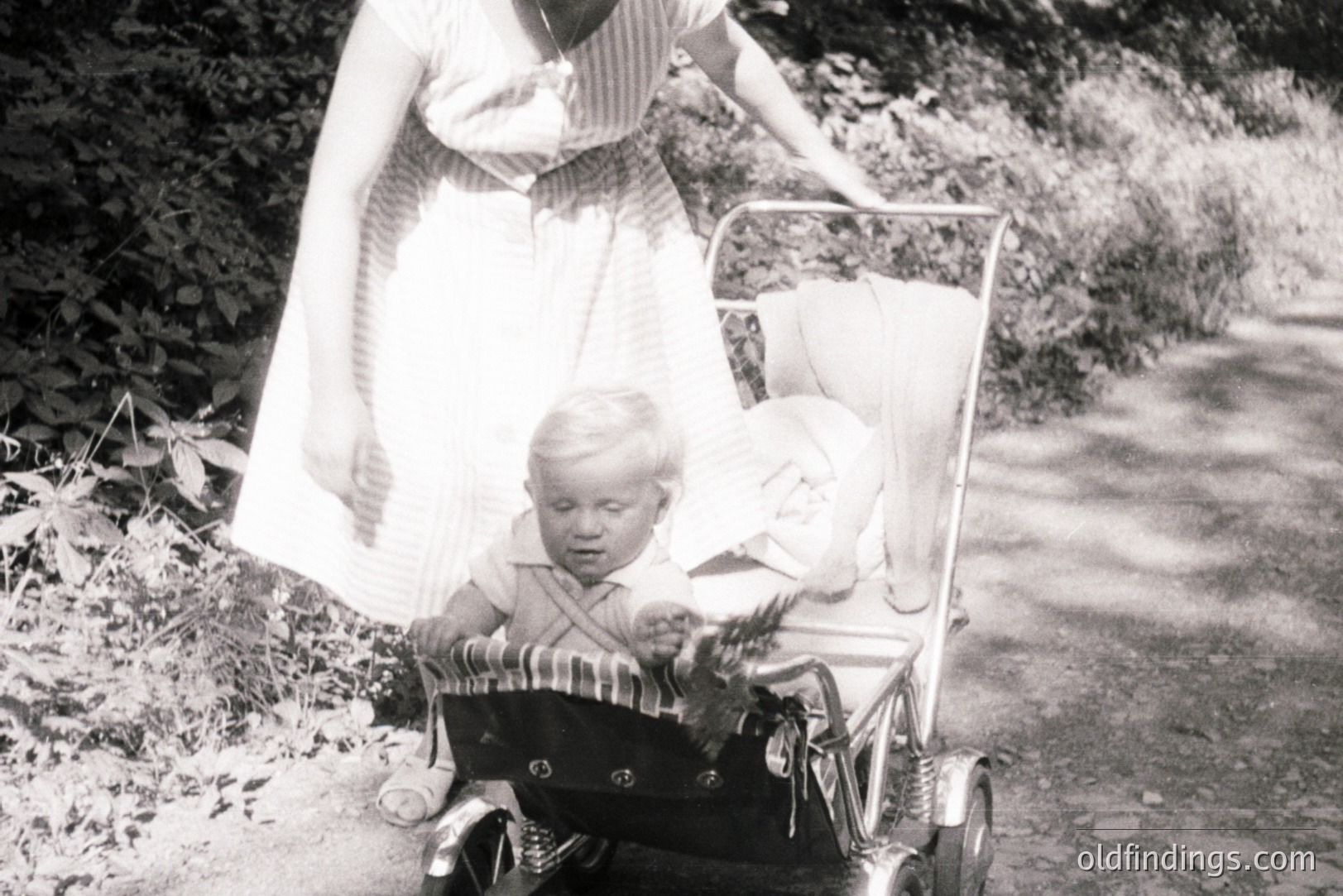 A young boy sits in a vintage, chrome-framed stroller, appearing slightly distressed. A woman in a patterned dress guides the stroller along a gravel path bordered by foliage. The scene conveys a mid-century domestic moment, possibly the 1960s. Excellent for design referencing or historical family studies.
