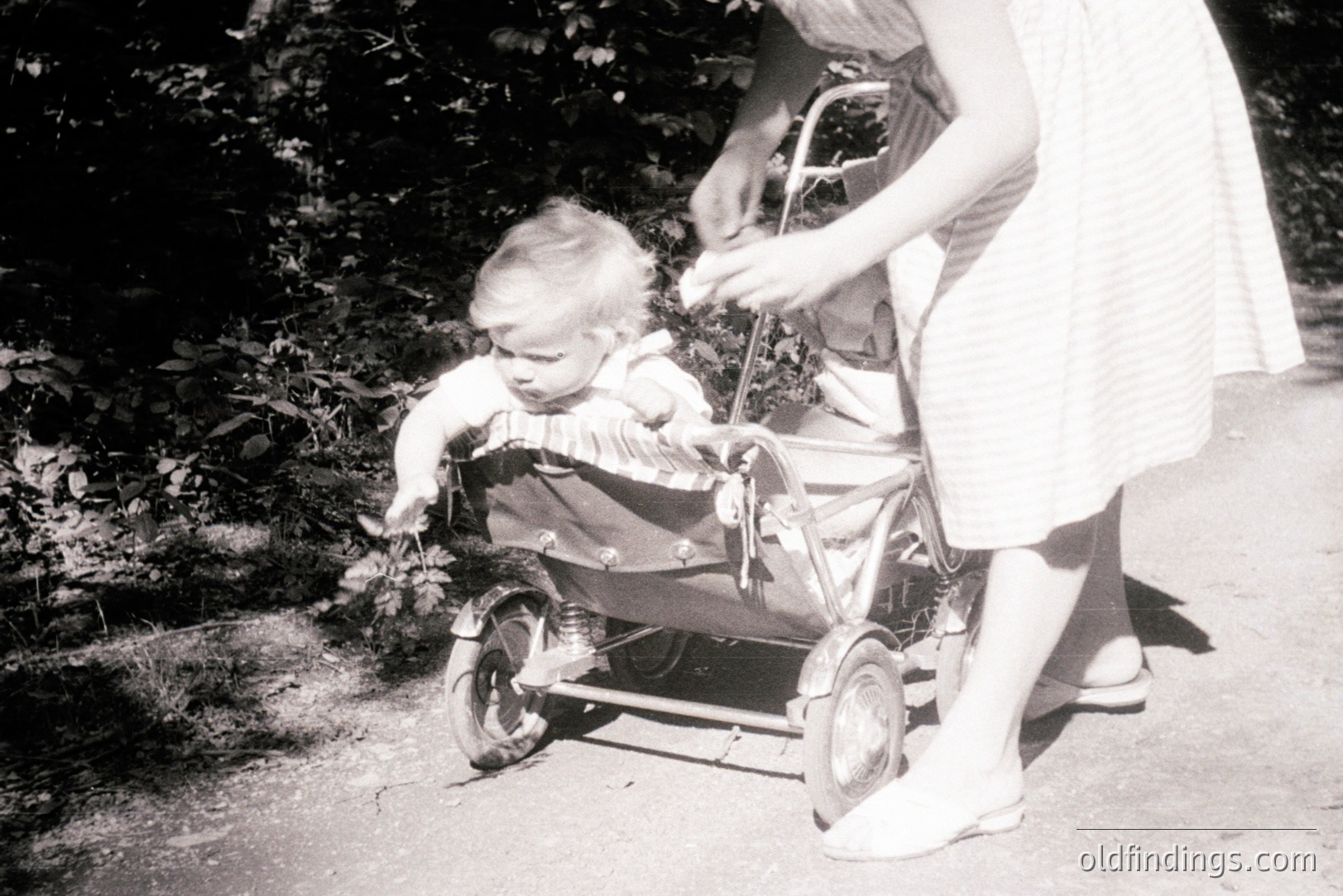 A black and white photo shows a young child seated in a vintage, metal baby carriage on a paved path. The child, wearing a short-sleeved garment, appears to be looking directly at the camera. A woman, partially visible, pushes the carriage from behind, wearing a striped dress and ballet flats. Likely 1960s domestic scene. Stock potential for retro design.