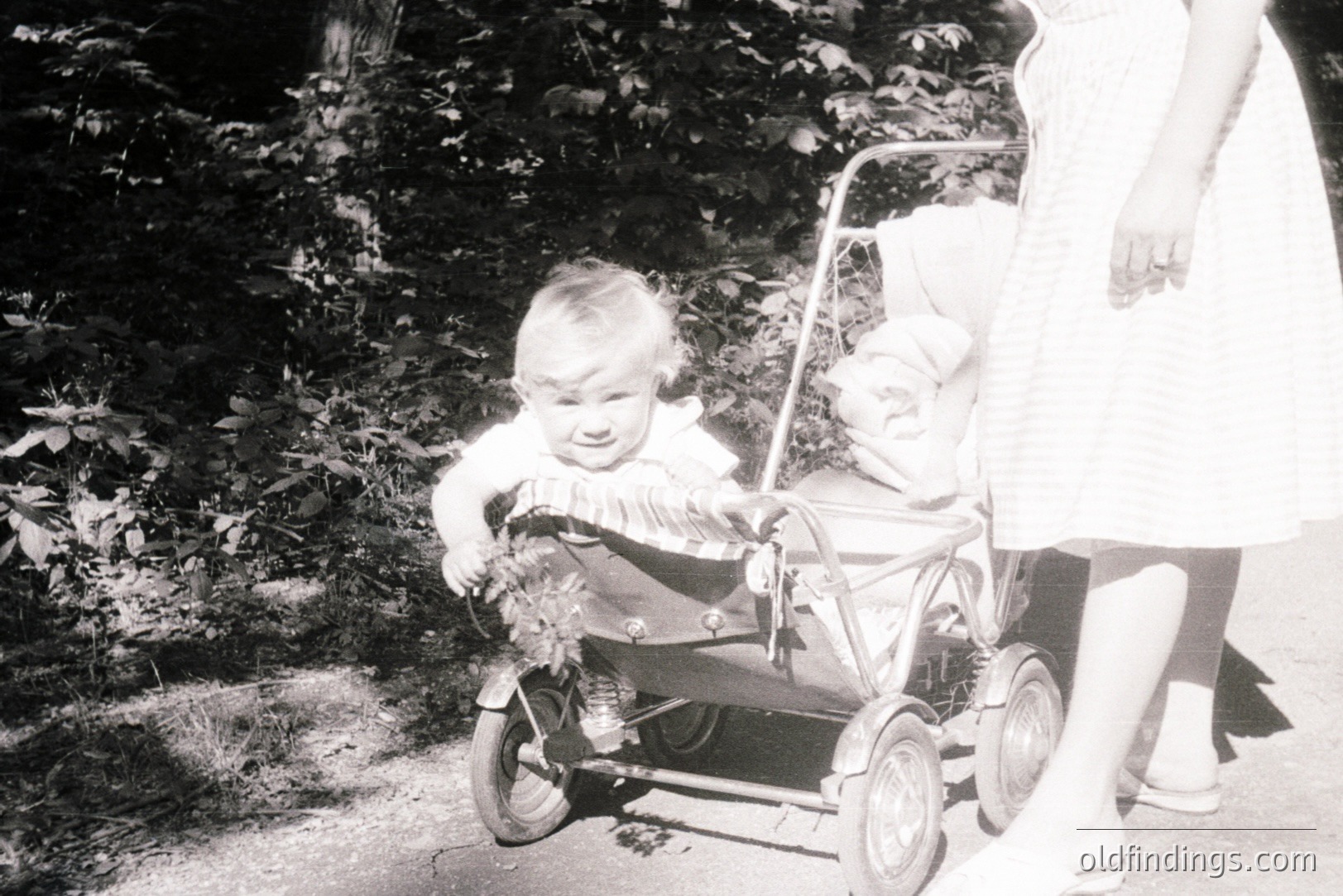 A young child sits in a vintage metal stroller, appearing to cry or express a strong emotion. A baby in a bonnet rests within the stroller’s upper compartment. The stroller is pushed by an unseen adult. Likely a candid family snapshot, circa 1950s-1960s.