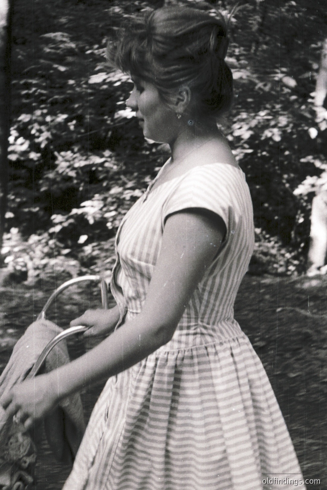 A young woman in a striped dress walks away from the camera, carrying a handled basket. Likely a candid snapshot, exhibiting 1960s fashion and photographic style. Soft focus and natural lighting suggest an outdoor setting, possibly a garden. Commercial value for design or vintage fashion references.