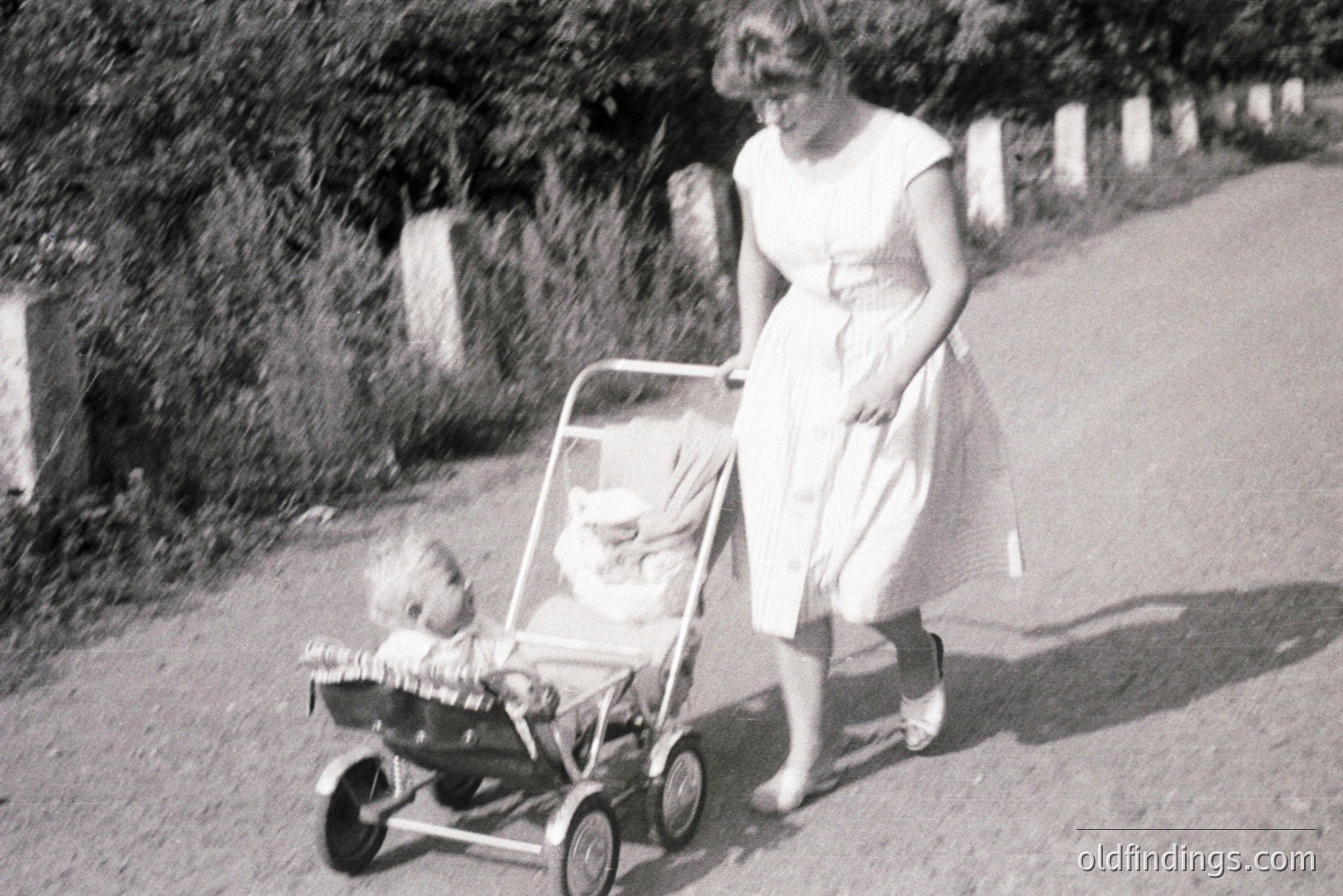 A woman in a light-colored dress pushes a baby in a vintage pram along a gravel road, framed by a stone wall and lush foliage. The scene evokes a 1960s domestic setting. Likely a family outing, captured with a snapshot aesthetic. Good potential for vintage design reference.