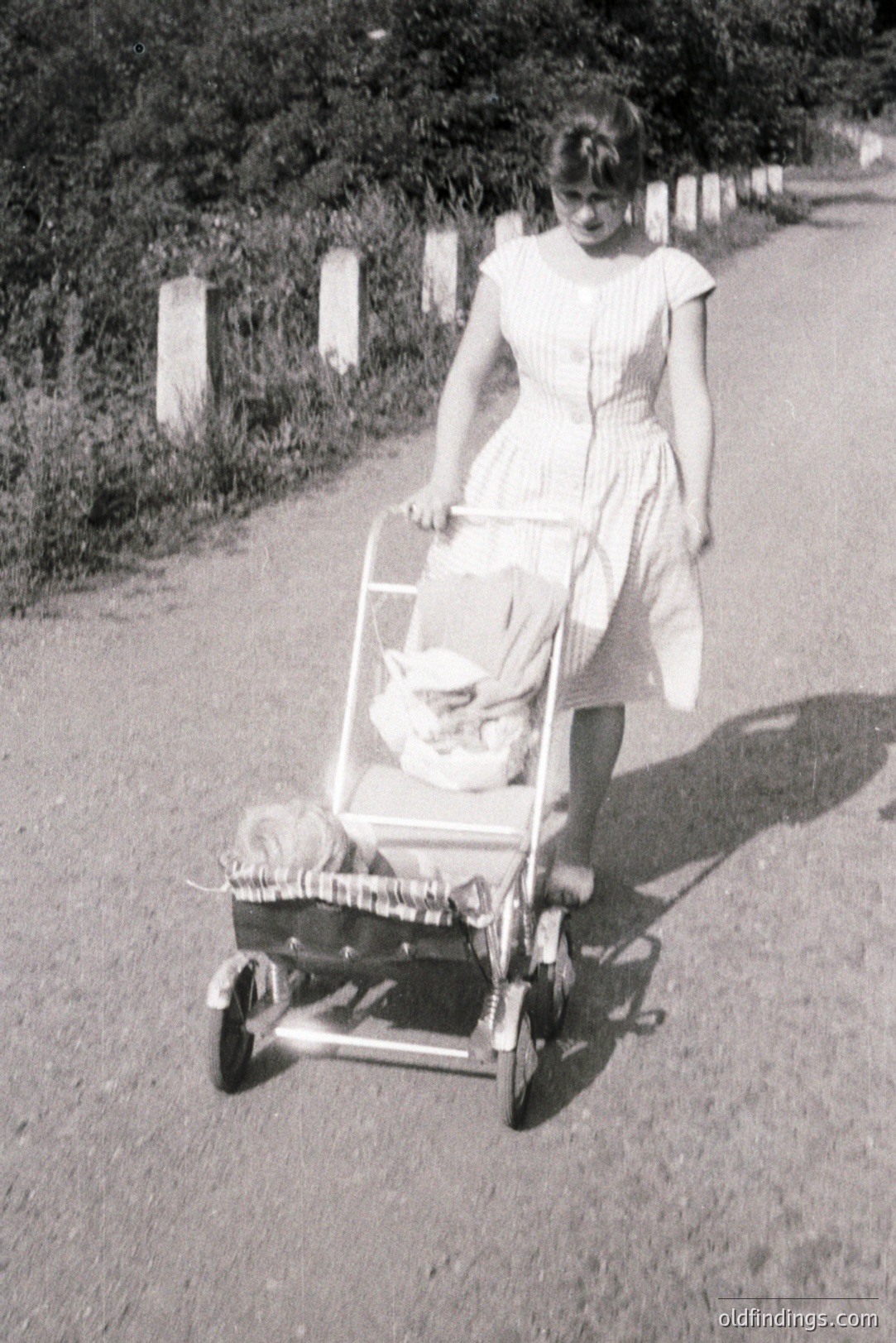 A young woman in a light-colored, patterned dress pushes a vintage silver baby carriage along a gravel road. The carriage is intricately detailed, showing craftsmanship. Appears to be mid-1960s, possibly a family snapshot. Historic domestic life.