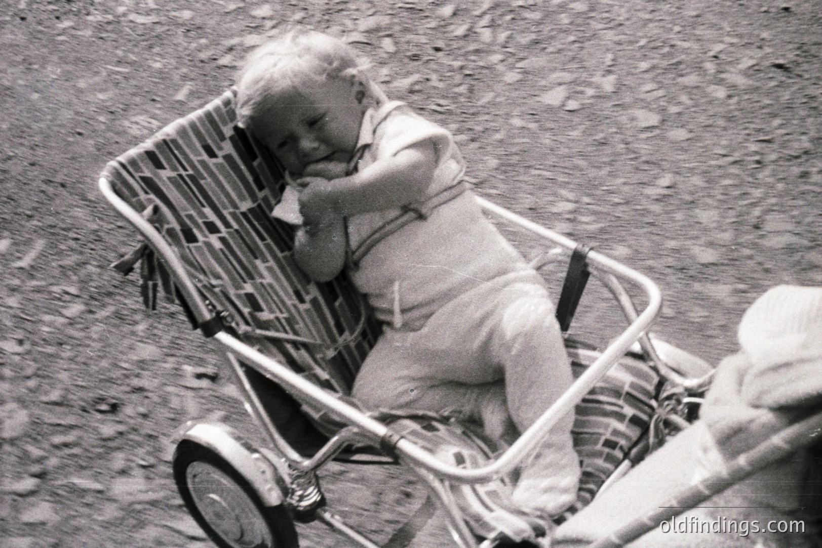 A young child rests in a wicker-framed stroller with padded seat, wearing a short-sleeved romper. The image, likely from the 1960s or 70s, shows a candid moment—the child appears sleepy or contemplative. The stroller’s design is typical of mid-century baby carriages.