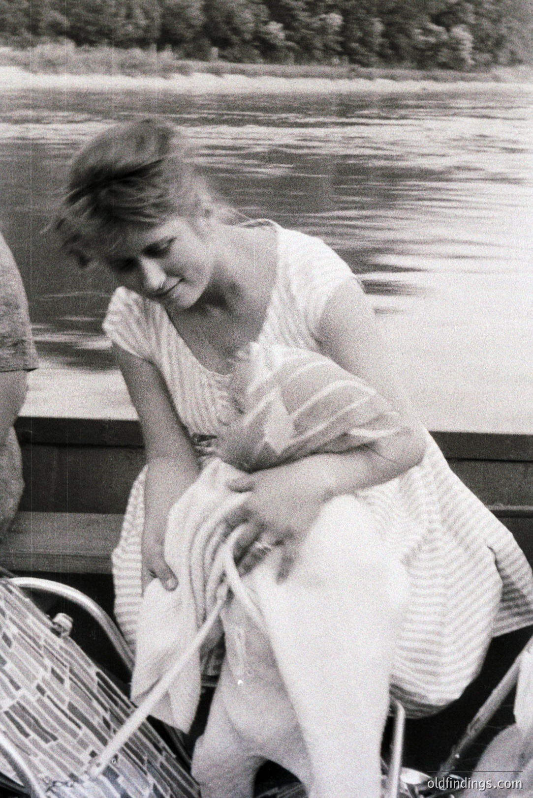 A young woman, seated on what appears to be a boat, gazes across a body of water. She wears a vertical-striped dress and holds a folded towel. Shallow depth of field and grainy texture suggest a candid moment, likely 1970s. Rustic boating aesthetic.