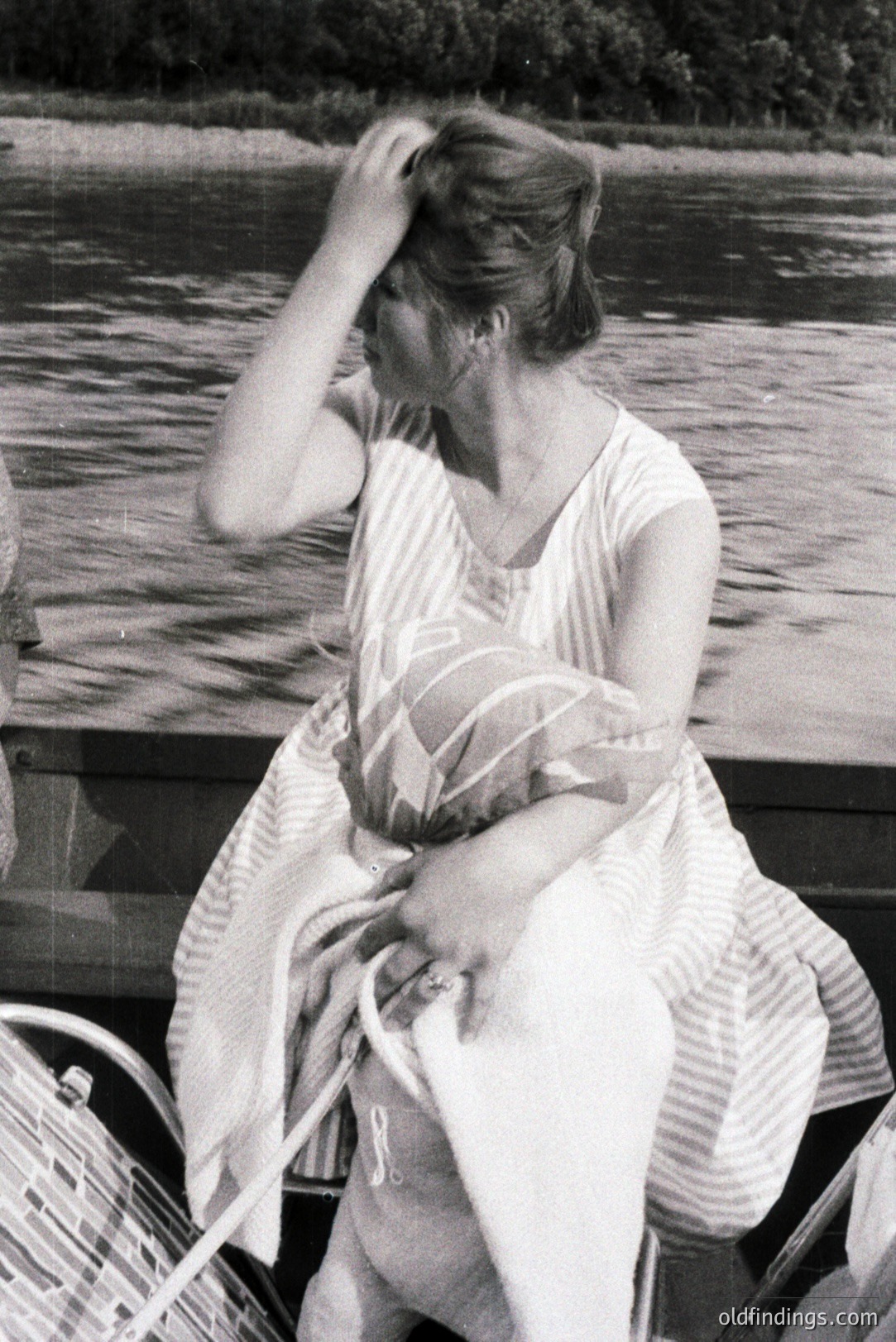 A woman in a striped dress sits outdoors, seemingly on a boat, with a blurred lakeside view. She appears to be pushing her hair back. Style suggests 1960s or 1970s fashion. Fine details are softened due to the image quality. Appears suitable for design or vintage aesthetic projects.