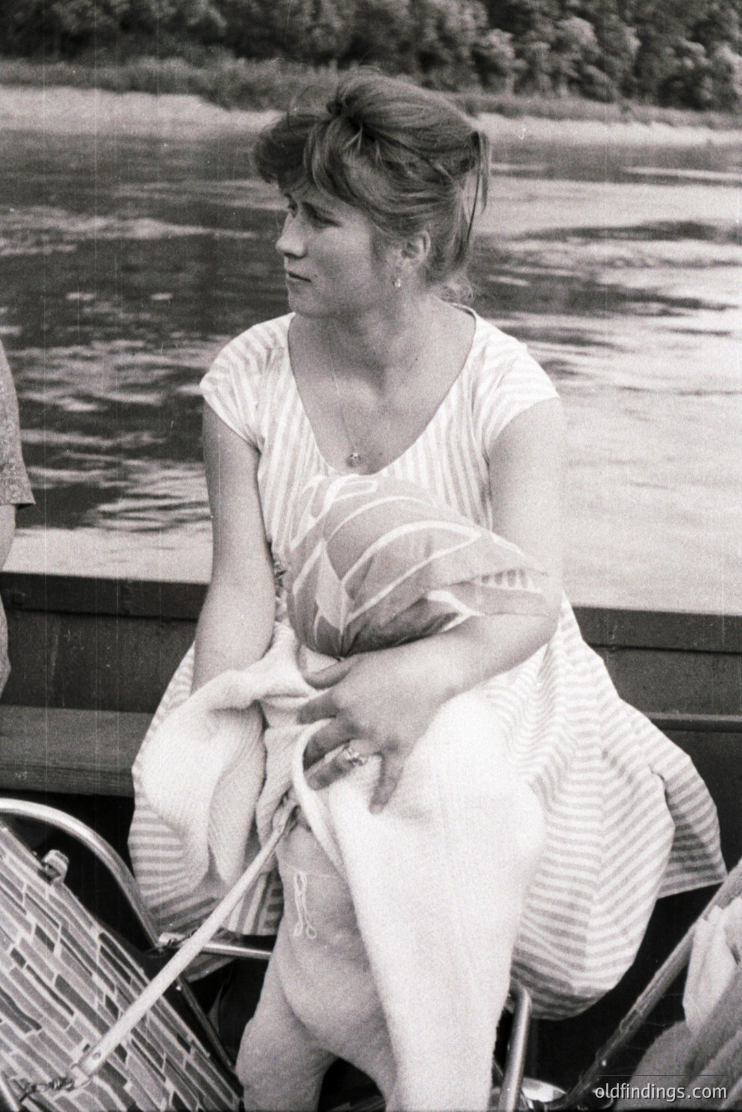 A woman in a striped dress sits cradling an infant, likely on a boat. She wears a delicate necklace and a ring. The background shows water and shoreline vegetation. Appears to be a casual, candid portrait, c. 1970s. Likely family documentation or a lifestyle stock photo.