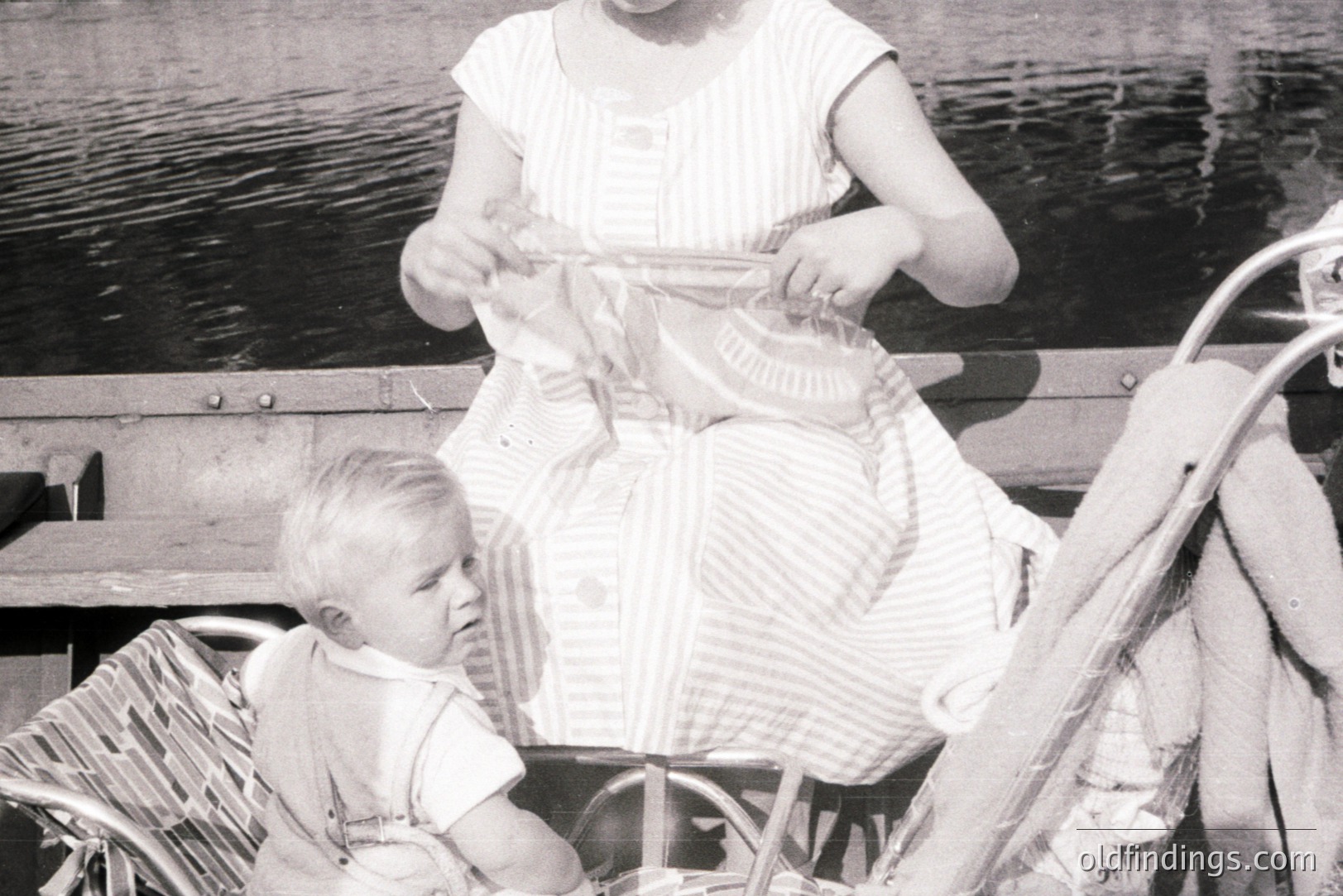 A young girl in a striped dress sits in a wicker stroller, focused on a cloth doll or toy. A toddler sits in the stroller's lower compartment, looking towards the camera. Likely a family portrait capturing everyday life. Appears to be a mid-century photograph, c. 1950s-1960s.