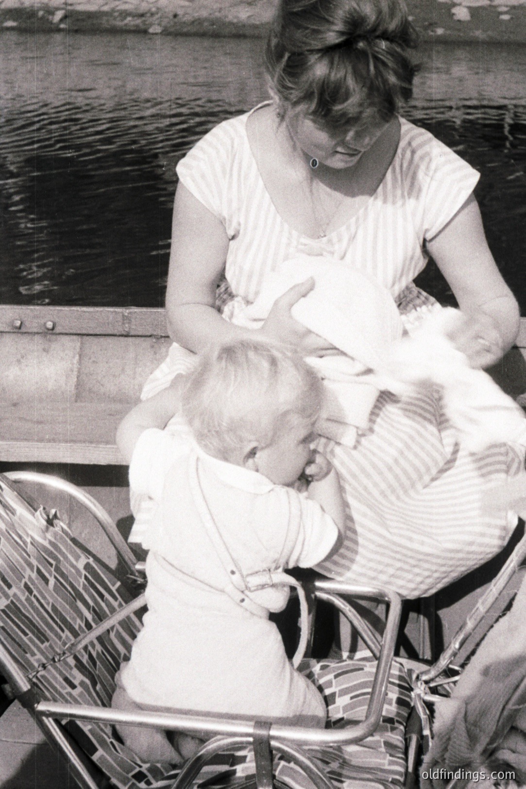 A mother tenderly soothes a baby in a high-backed, striped pram. The scene suggests a park setting, with blurred foliage visible in the background. The mother's simple dress and the pram's design indicate a likely timeframe of the 1960s or 1970s. Intimate and evocative.