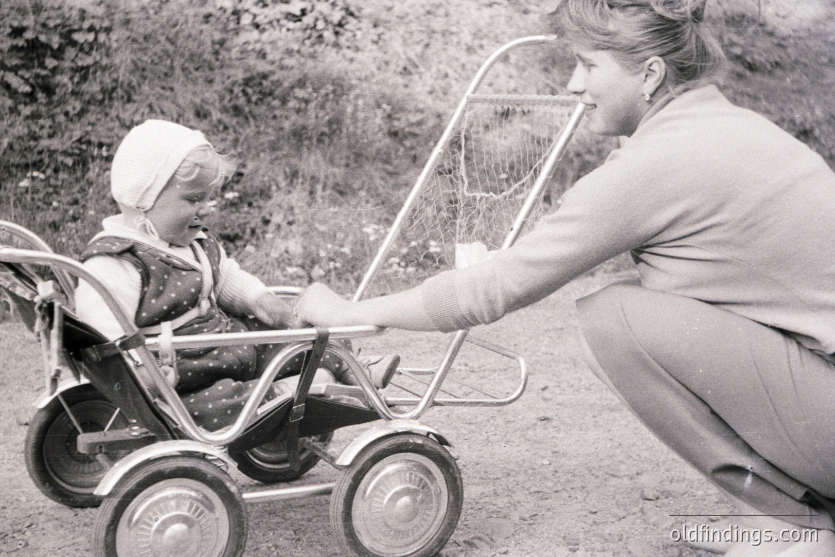 A woman kneels beside a young child seated in a vintage baby carriage with a wire basket. Child wears a bonnet and patterned dress. The scene evokes a candid, intimate family moment. Likely taken in the 1960s or 70s, capturing everyday domesticity. Valuable for nostalgia and design reference.