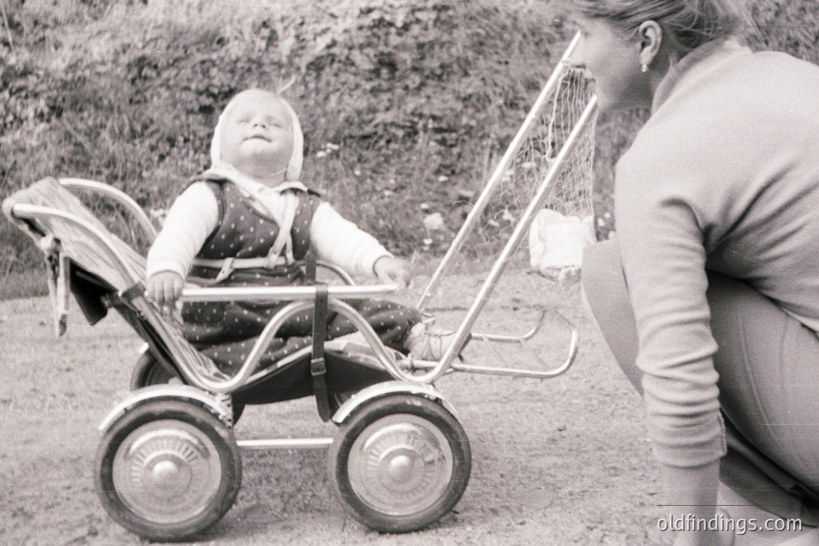 A young child sits serenely in a vintage, chrome-framed stroller. A woman kneels nearby, offering a gentle interaction. The stroller's design suggests a mid-century aesthetic, possibly 1950s or 60s. Location is outdoors, on a gravelly surface. Potential stock photo for vintage parenting themes.