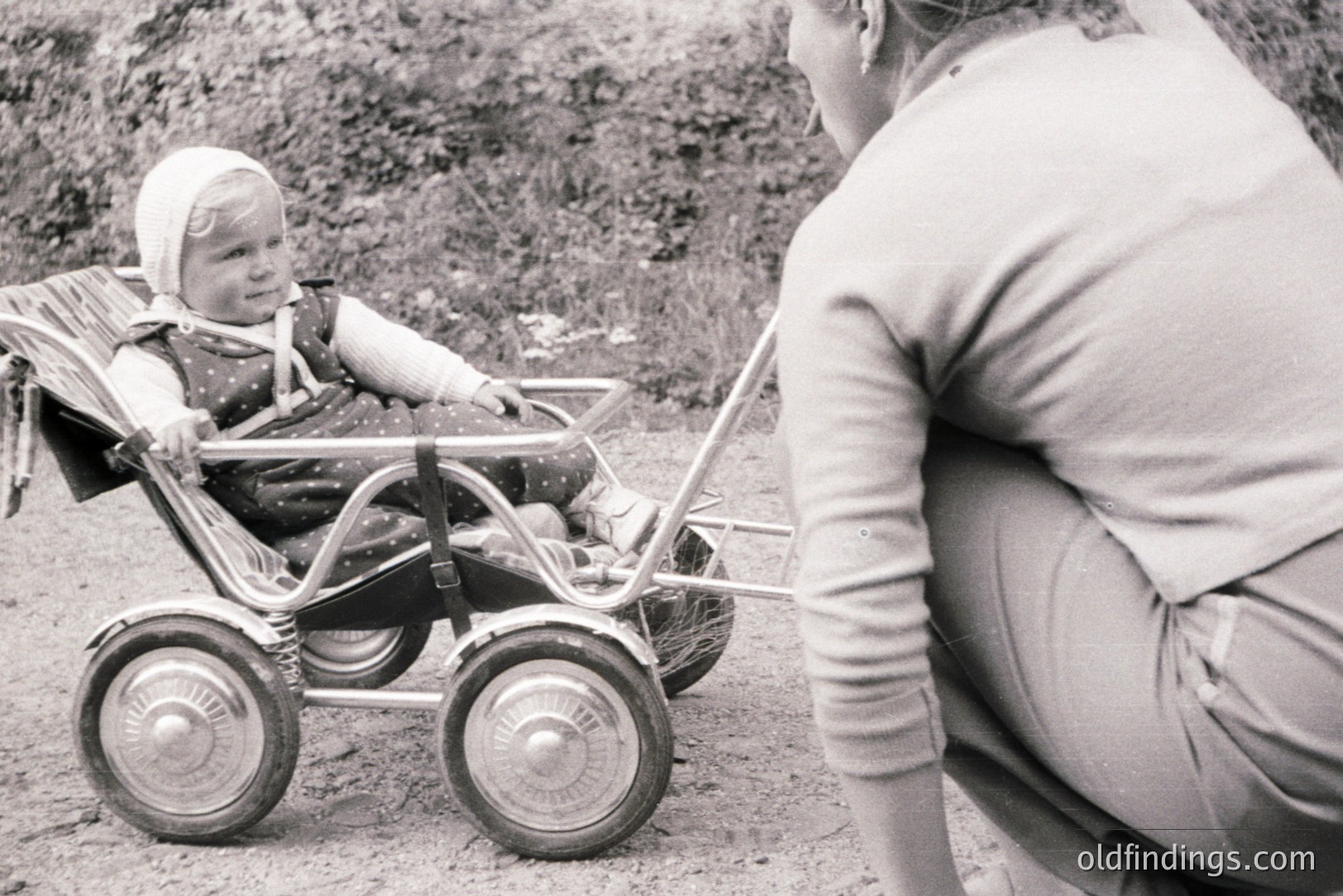 A toddler sits in a vintage, metal doll stroller, pulled by a caregiver in a long-sleeved sweater. The scene depicts a domestic moment, likely outdoors on a grassy or dirt patch. Appears to be a candid, snapshot-style photograph. Estimated timeframe: 1960s-1970s. May serve as a reference for vintage toy or childhood design.