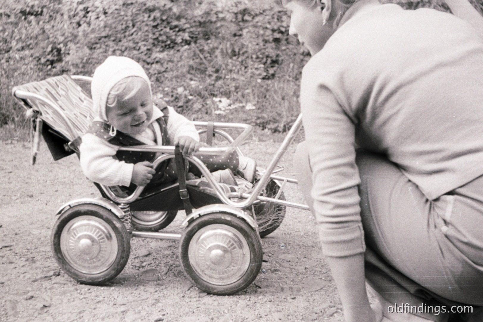 A toddler in a vintage metal stroller, wearing a bonnet and overalls, grins. An adult, possibly a caregiver, is crouched nearby. The stroller's design suggests a 1950s-60s timeframe. Appears to be an outdoor, possibly rural, setting. A charming, nostalgic family moment captured.