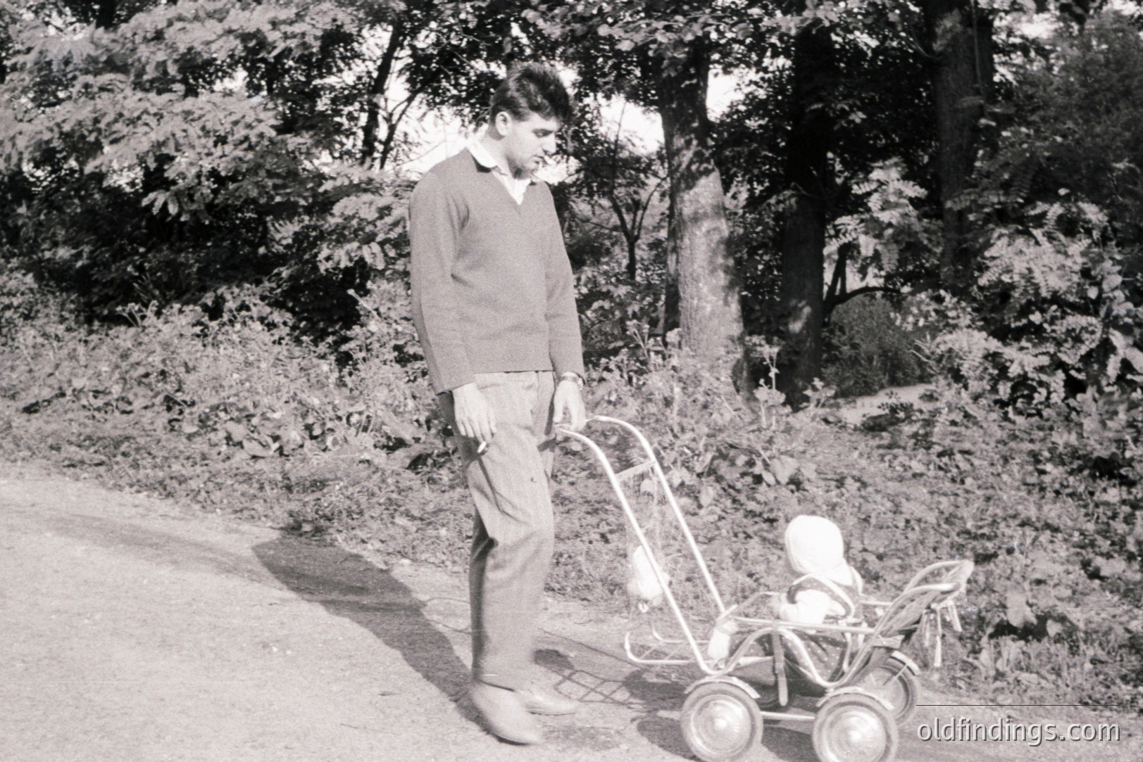 A young man in a sweater and trousers strolls along a gravel road, pushing a vintage doll carriage. Likely mid-20th century, possibly 1950s-1960s. Soft lighting & grainy texture characteristic of film photography. Image evokes nostalgia and domestic life. Ideal for design or historical research.
