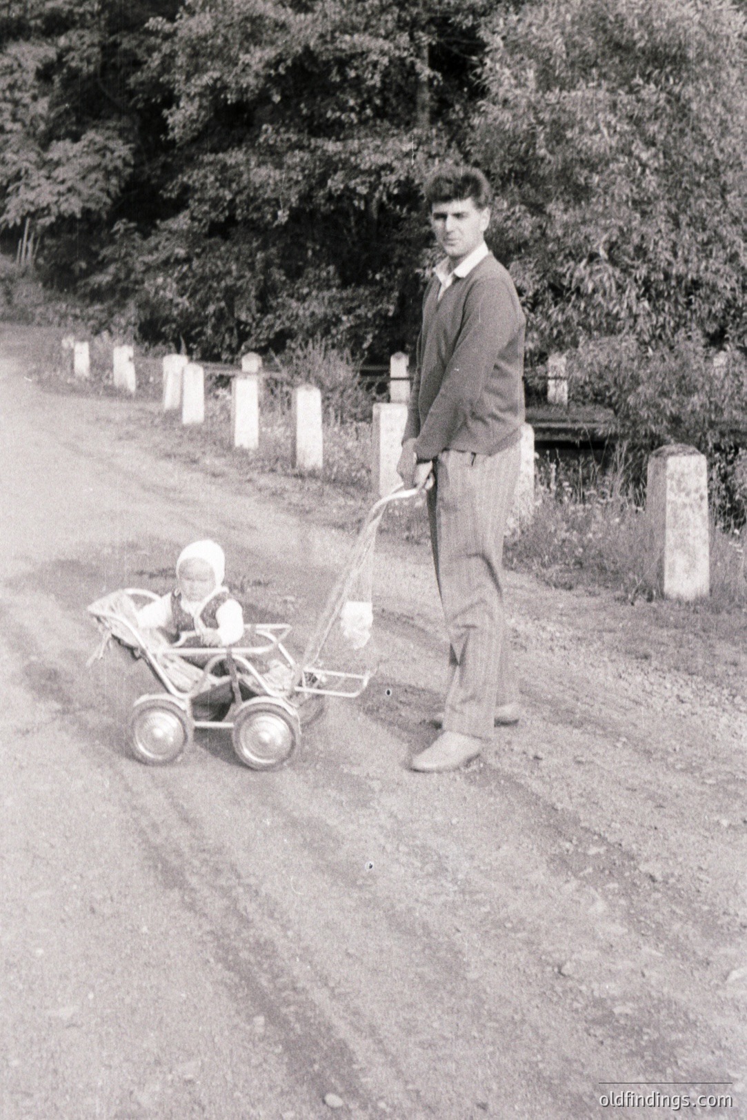 A young man in a tweed sweater and trousers stands beside a vintage stroller with a baby bundled within. The gravel road and stone wall suggest a rural setting. Likely a family snapshot from the 1960s or 70s, capturing a quiet moment. The stroller’s design indicates a mid-century aesthetic.