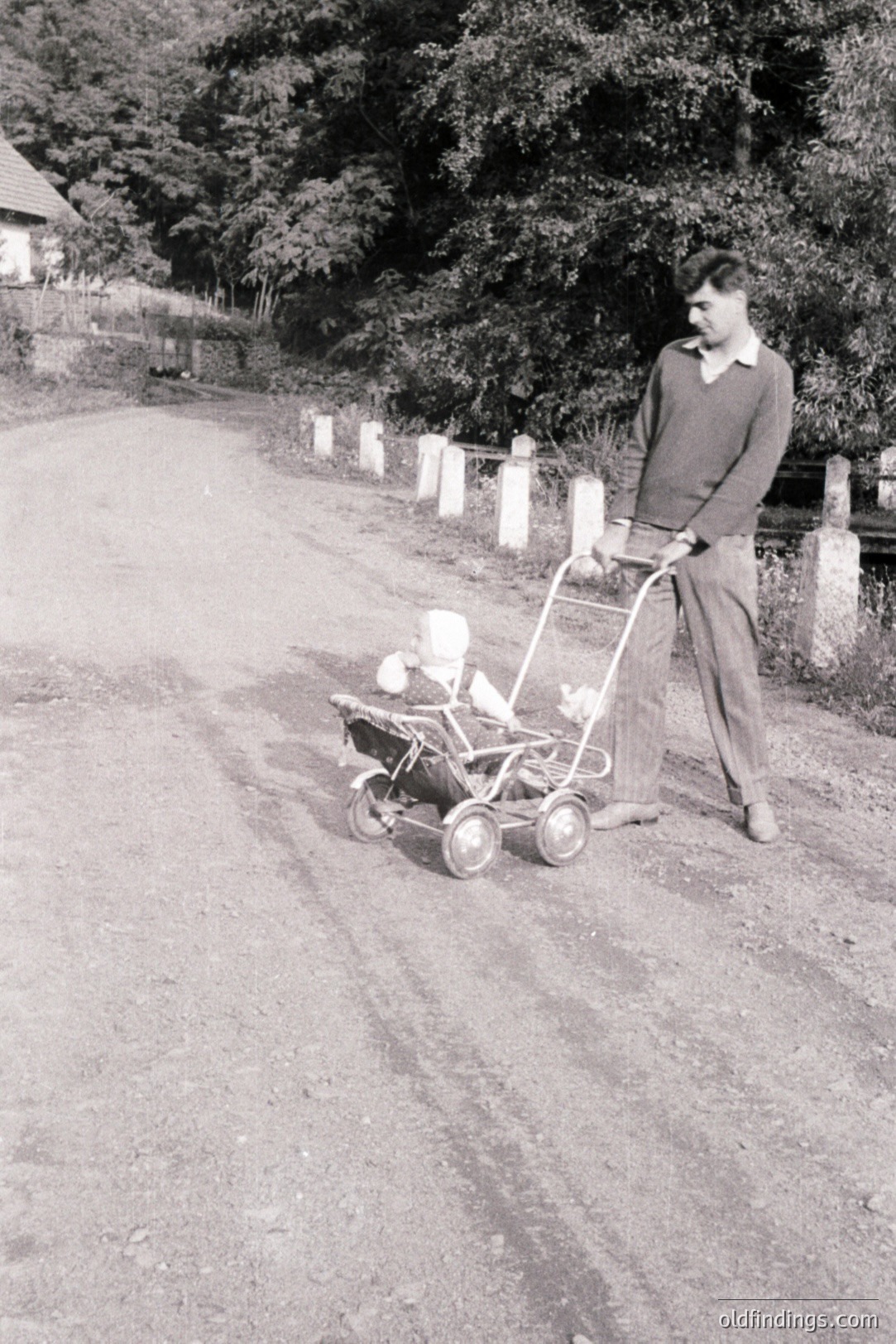 A man in a cable-knit sweater and trousers pushes a vintage rotary lawnmower along a rural, gravel road. Stone pillars line the roadside, with dense foliage visible beyond. Likely taken in the mid-20th century, possibly 1950s-1960s, this image offers a glimpse of domestic life and early lawn care technology.