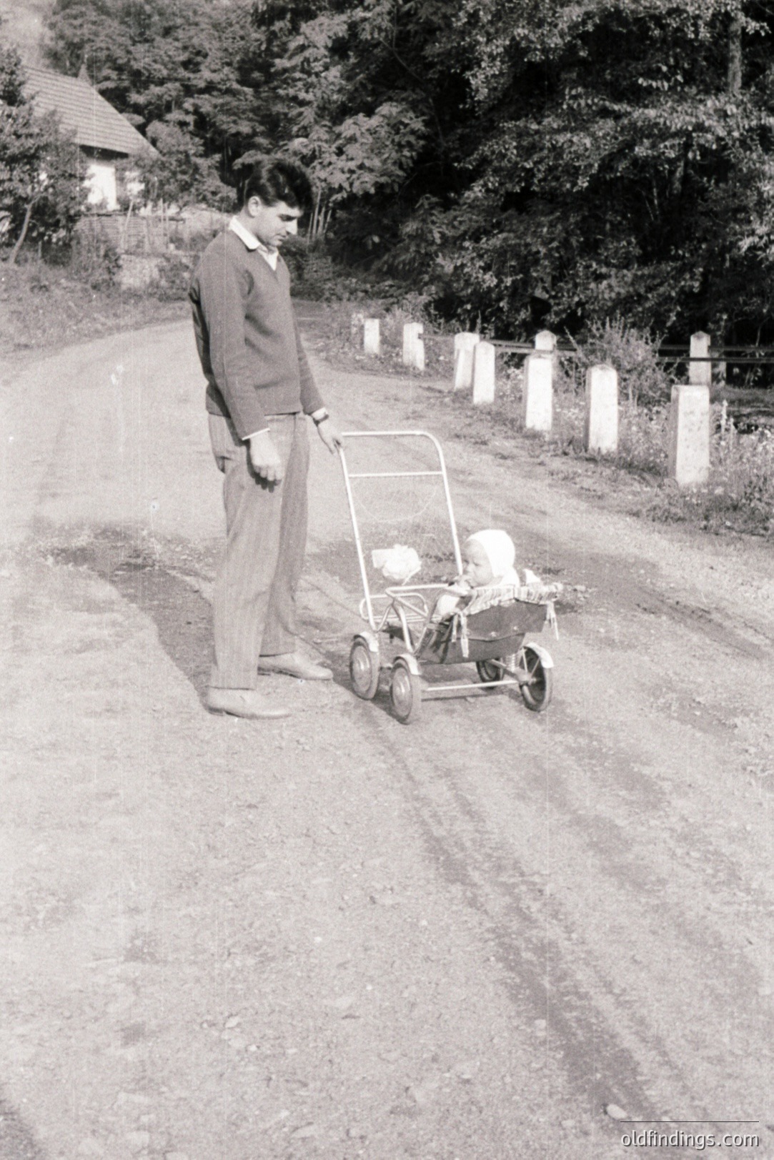 A young man in a sweater and trousers stands beside a vintage wicker baby carriage on a dirt road. Stone posts line the roadside, hinting at a rural location. The scene evokes a nostalgic, mid-century domesticity. Appears to be a candid moment, likely 1950s-1960s.