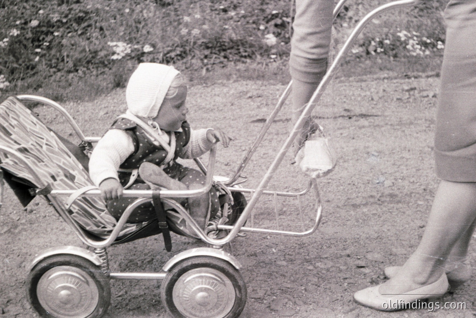 A young child, wearing a patterned dress and bonnet, sits in a vintage silver stroller, held by an adult. The stroller features shiny chrome details and black rubber tires. Likely 1960s, capturing a moment of childhood and domesticity. Appears to be an outdoor scene with greenery visible.