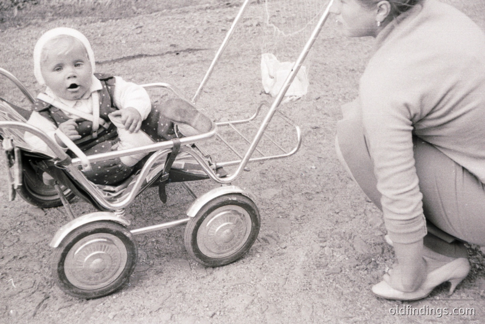 Black and white photograph shows a baby seated in a vintage, chrome-framed stroller. The baby wears a bonnet and formalwear. A woman in slacks and heels stands nearby, seemingly attending to the child. Likely a candid snapshot from the 1960s or 1970s, capturing a domestic moment.